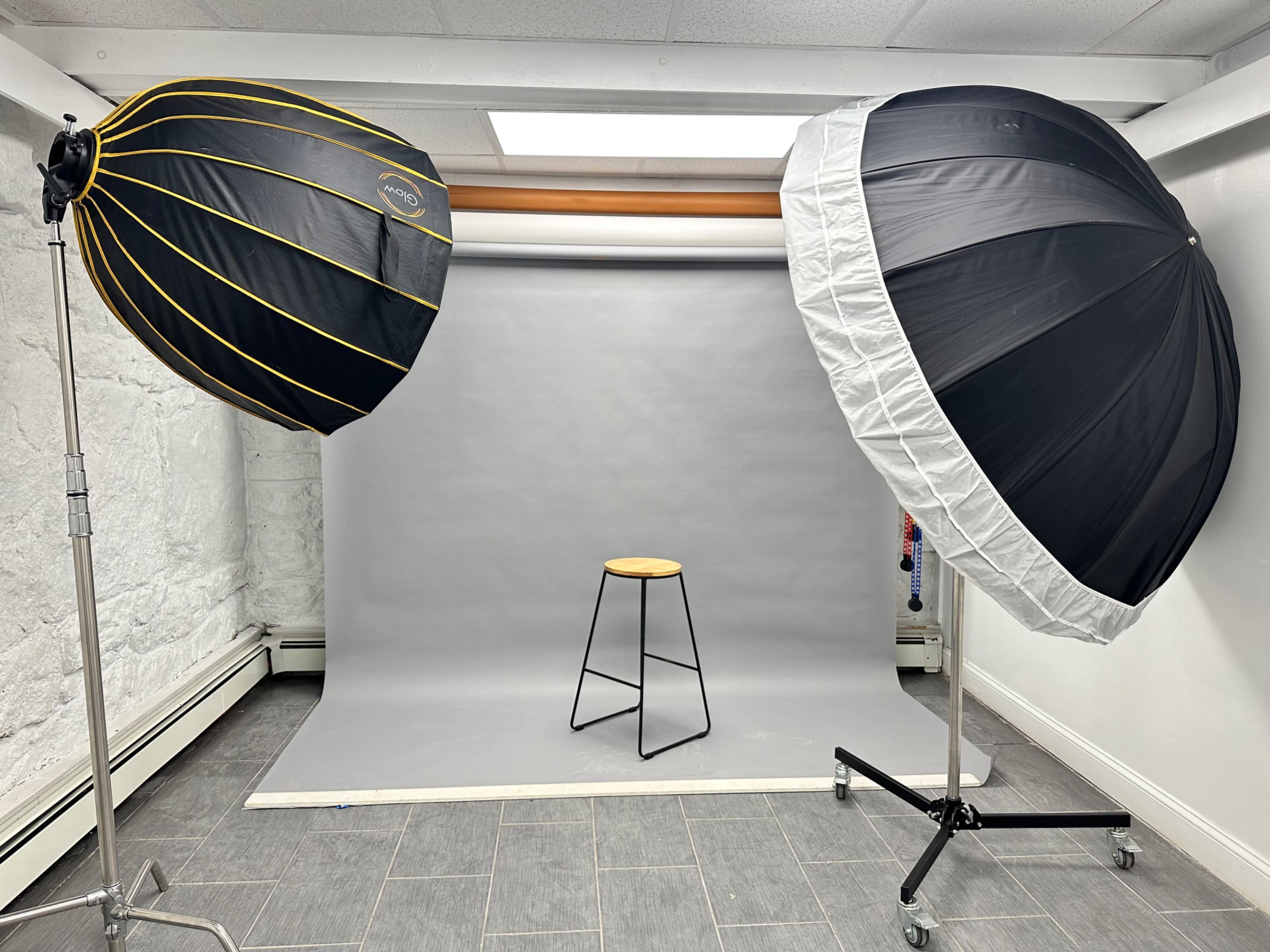 The image shows a photo studio setup with two large umbrella softboxes positioned on either side of a plain gray backdrop and a wooden stool in the center.