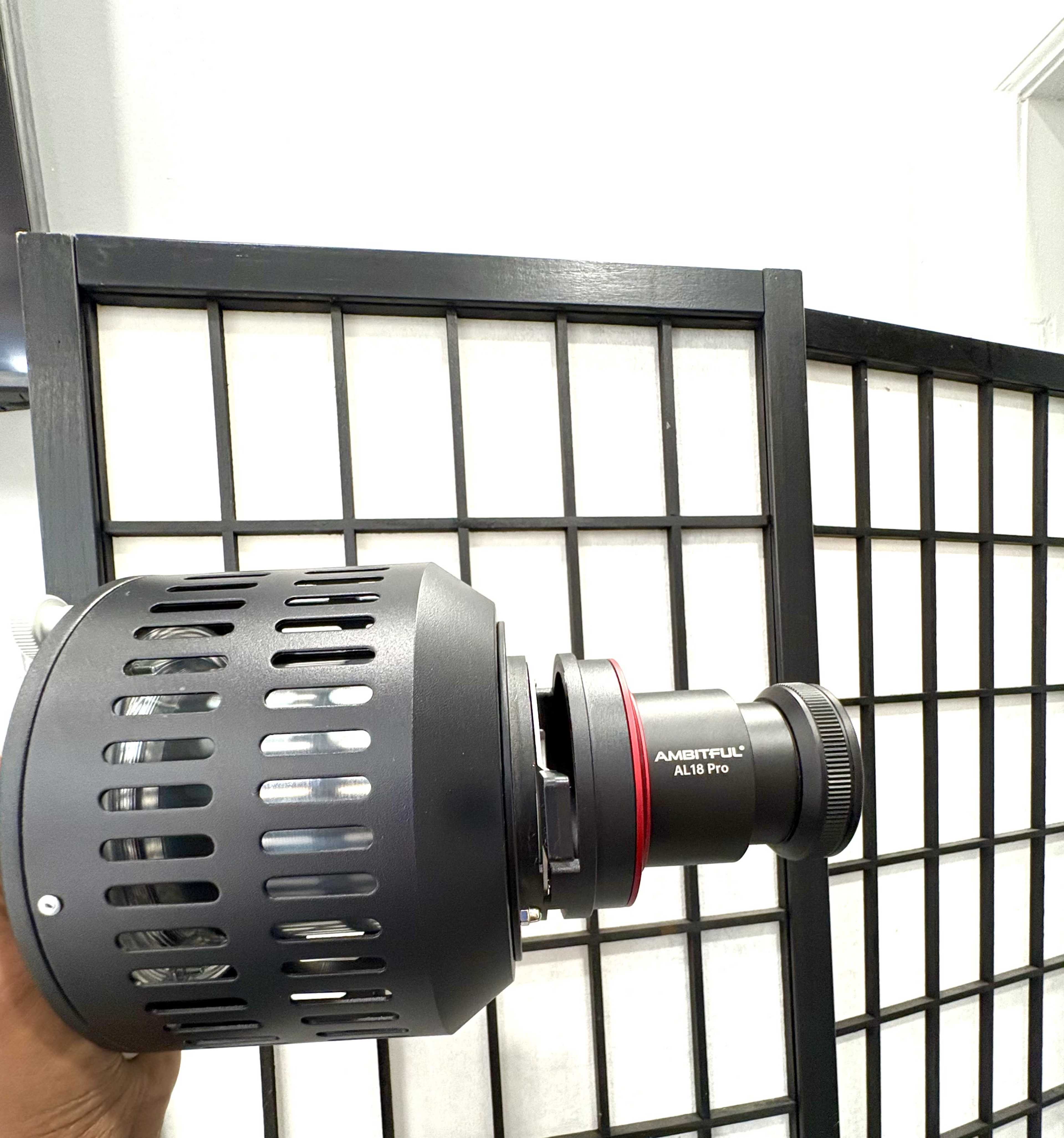 A hand holds a large black and silver camera lens in front of a partition with a grid pattern.