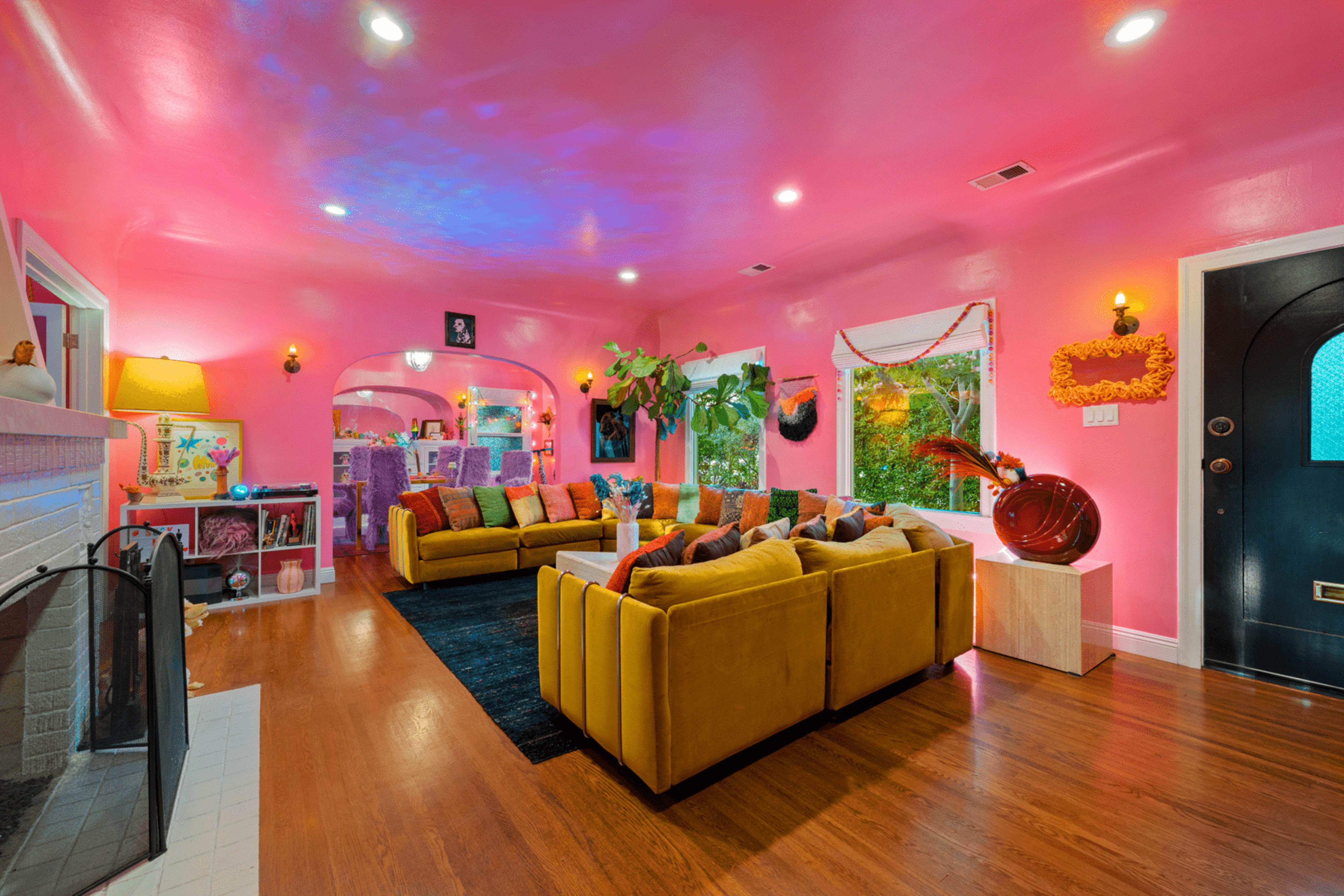 The living room features bright pink walls, a yellow sofa, and various plants beside a dark area rug and a fireplace.
