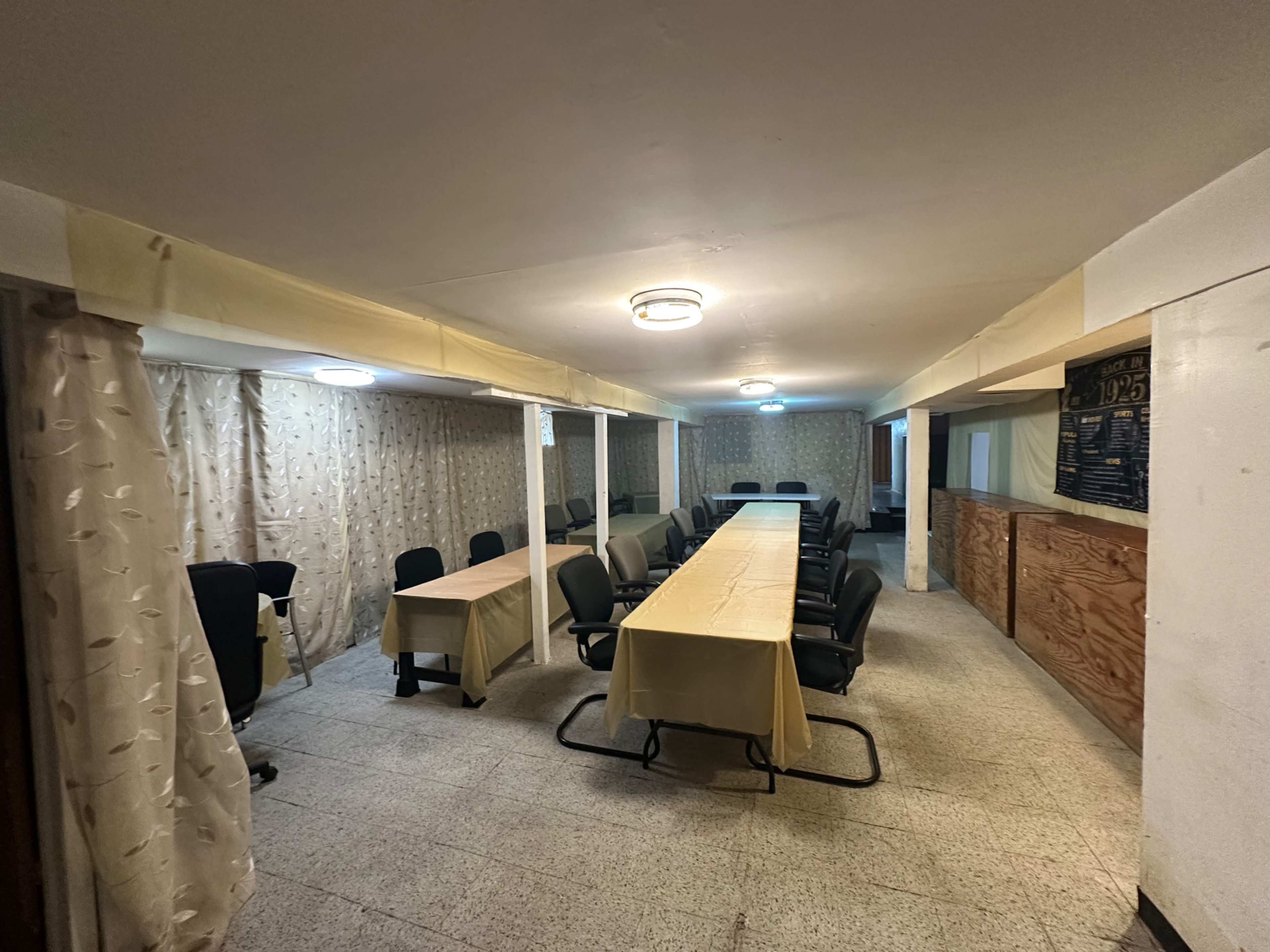 A rectangular room with multiple tables arranged in a long format, surrounded by chairs, and featuring light-colored curtains and wall panels.