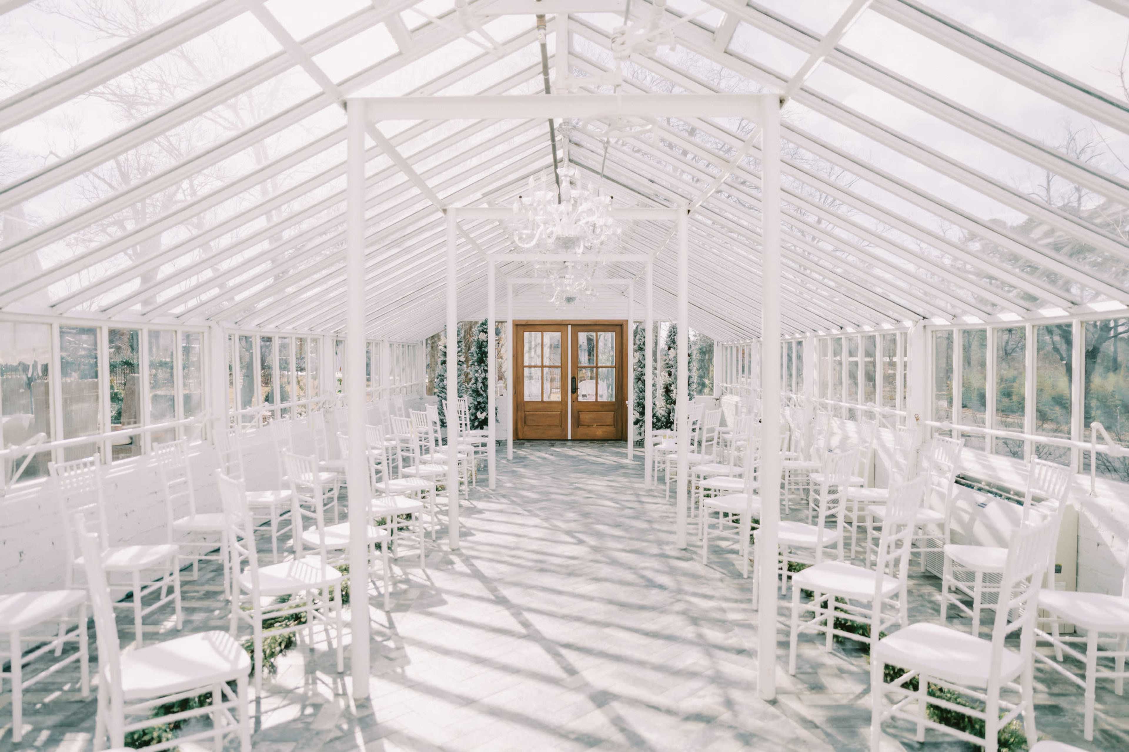 The image shows a bright, white glasshouse interior with rows of empty white chairs facing a set of double wooden doors.