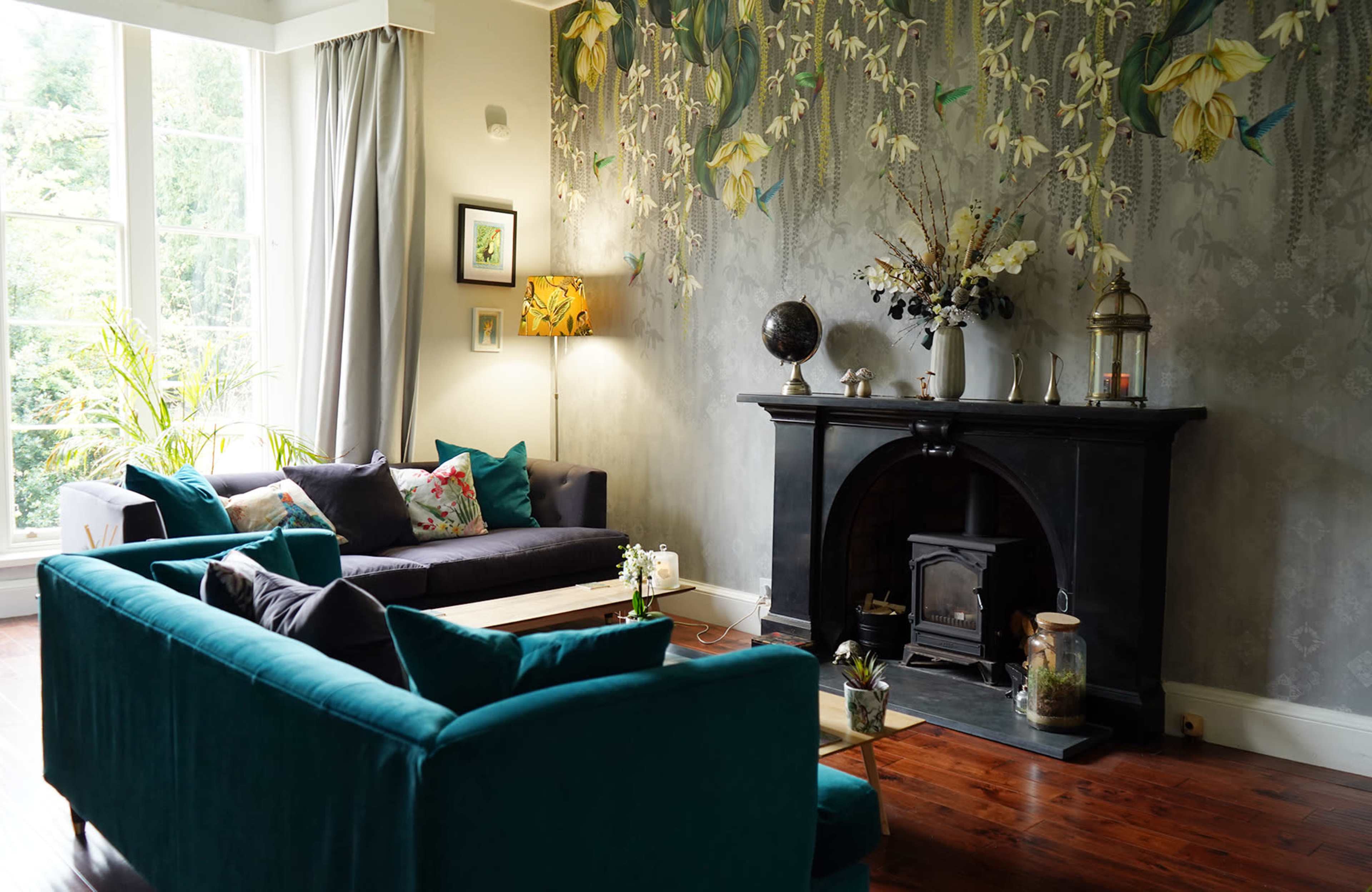 A living room features a dark fireplace, a large window with natural light, and a floral wall design, complemented by modern furniture in teal and dark hues.