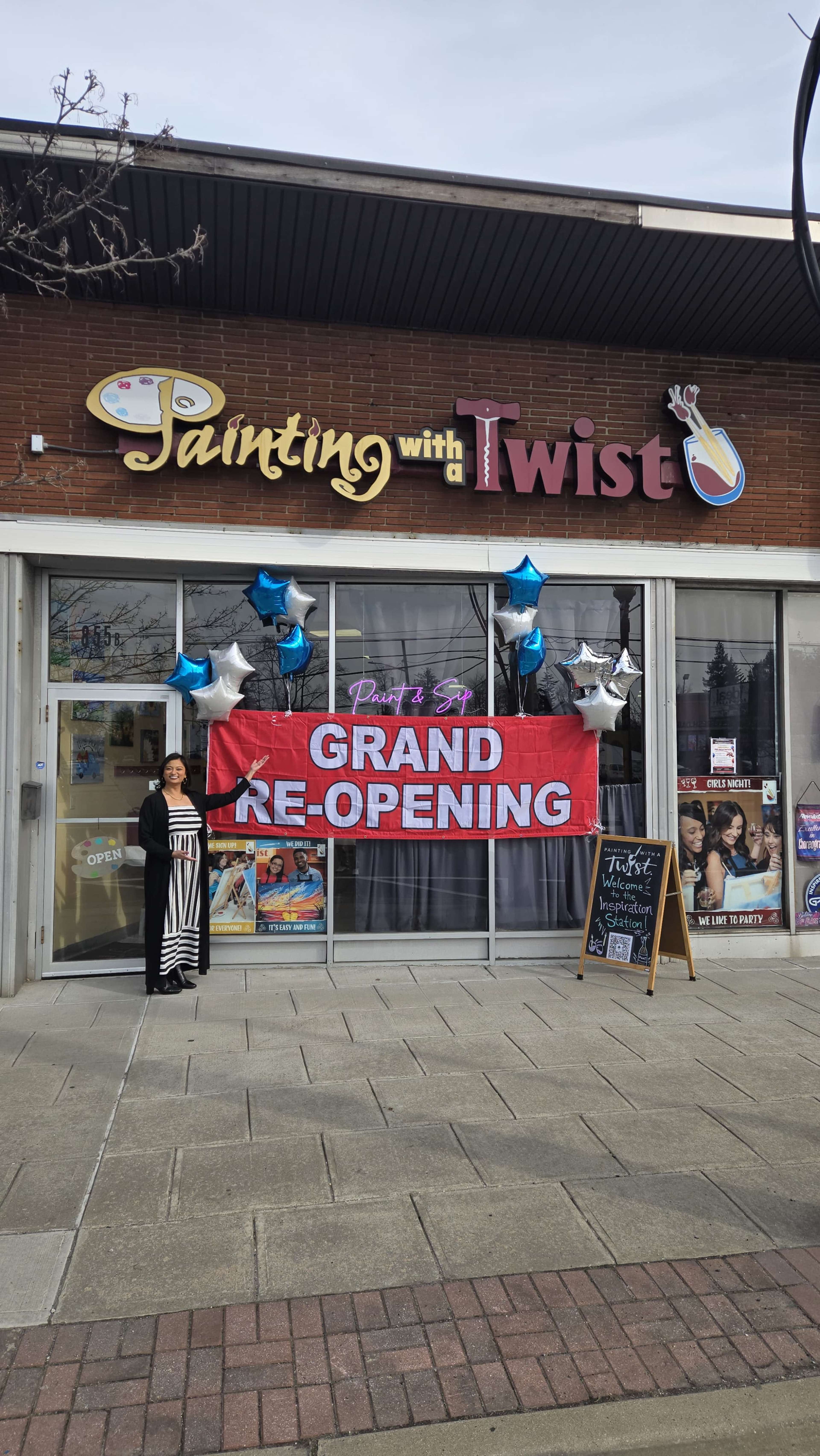 A woman stands in front of a storefront decorated with a large banner that reads "GRAND RE-OPENING," accompanied by blue and silver balloons.