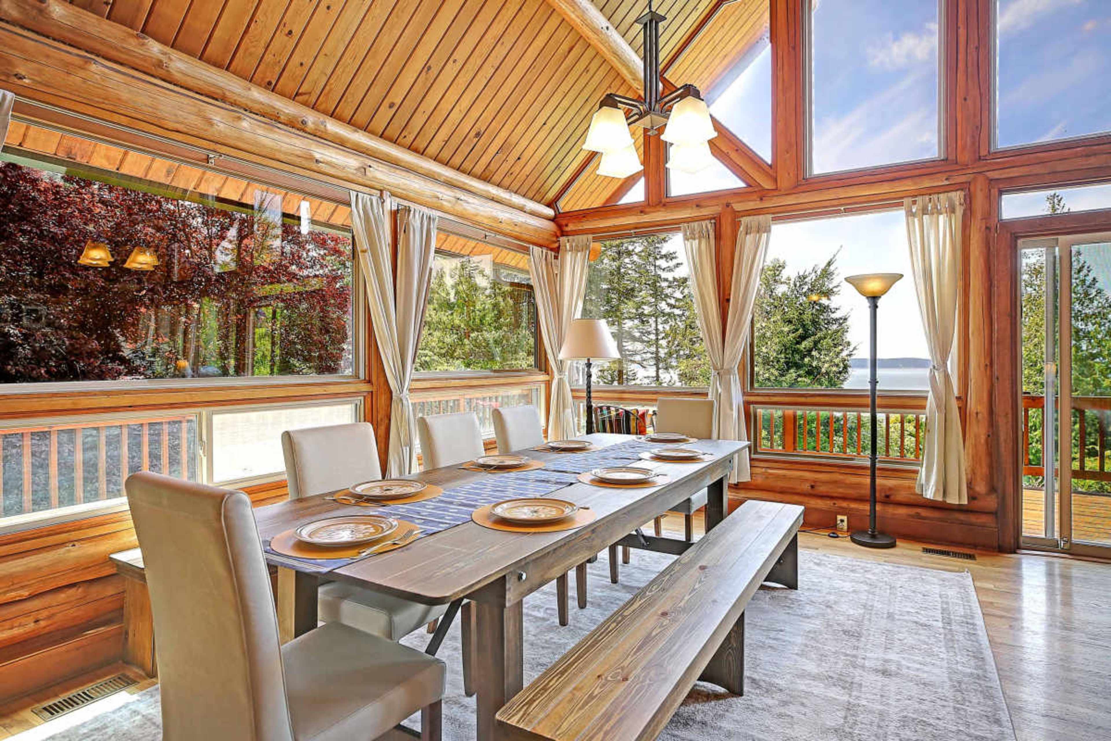 The image shows a dining area with a wooden table and benches, surrounded by large windows that offer a view of trees and water.