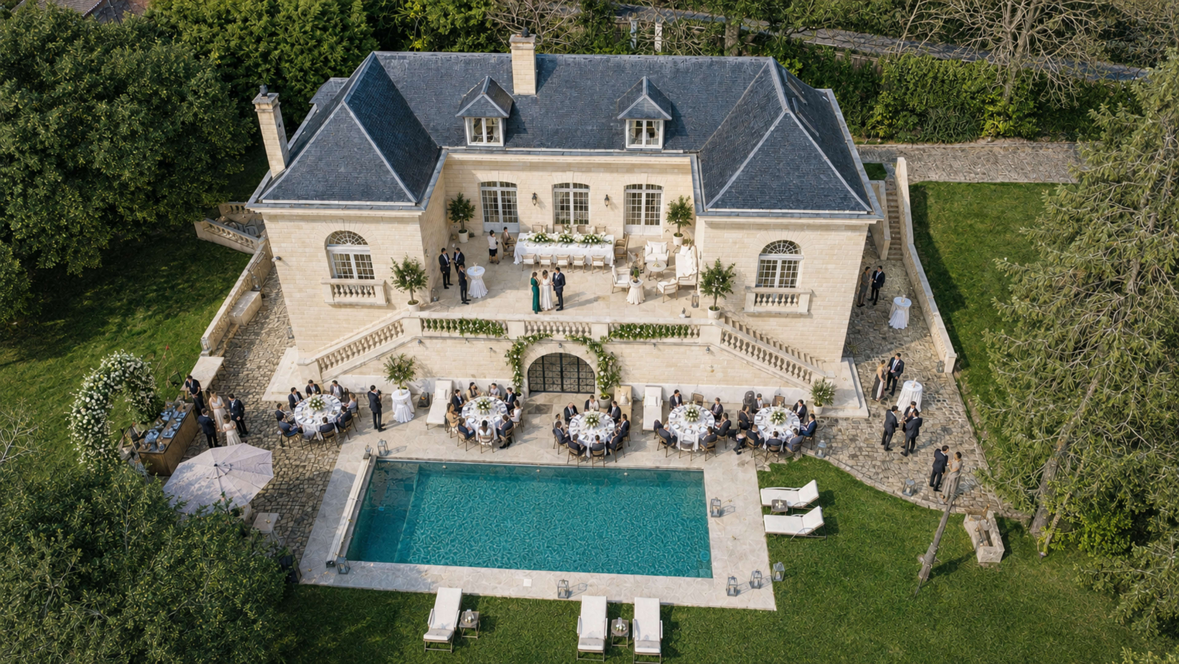 A large stone house with a dark roof overlooks a swimming pool and a gathering of elegantly dressed guests seated at tables in a landscaped garden.