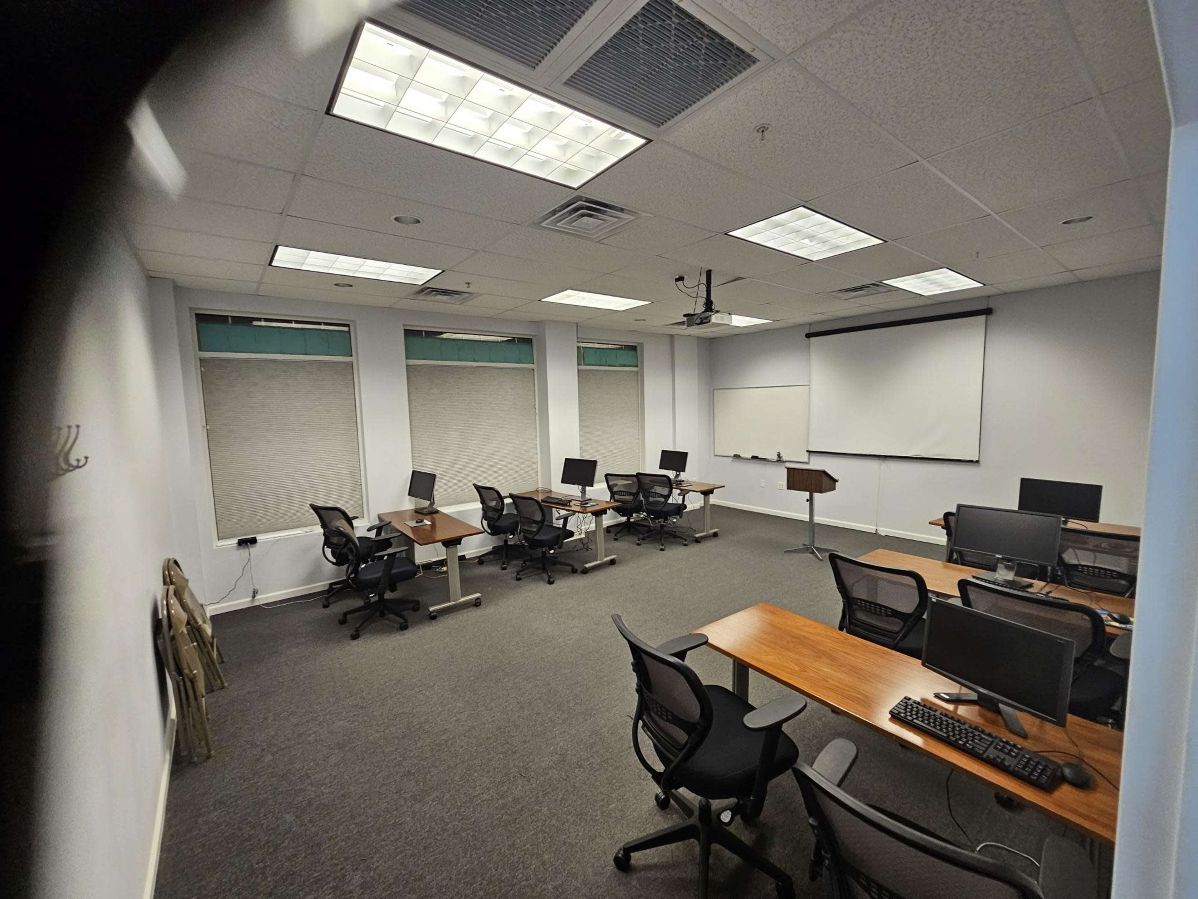 A spacious, empty conference room with several desks equipped with computers and a projector screen at the front.