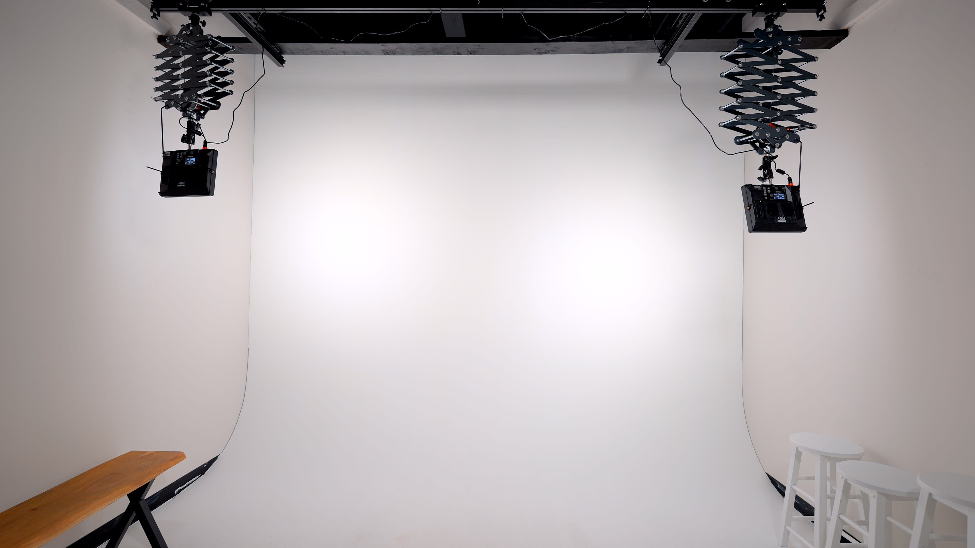 The image shows a photography studio with a plain white backdrop, two overhead lights, and a wooden bench alongside three white stools.