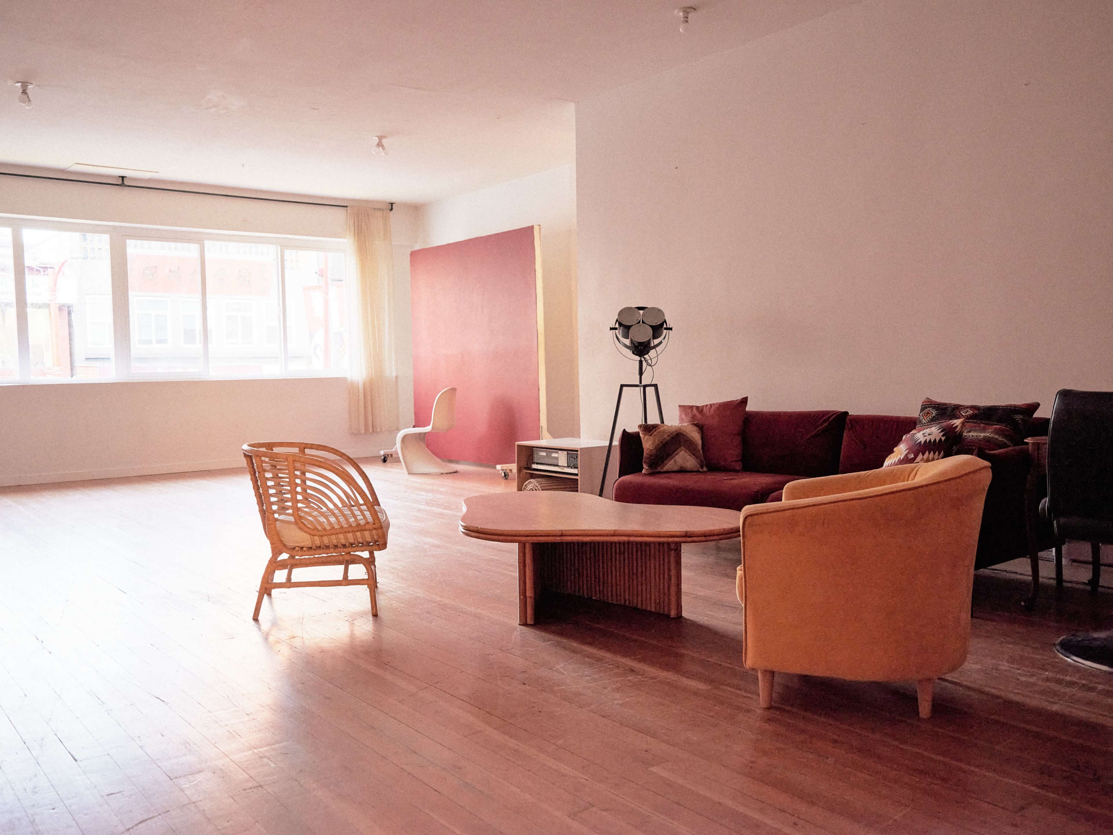 The image shows a spacious, minimally furnished interior featuring a red accent wall, a sofa, a light-colored chair, a wooden coffee table, and large windows providing natural light.