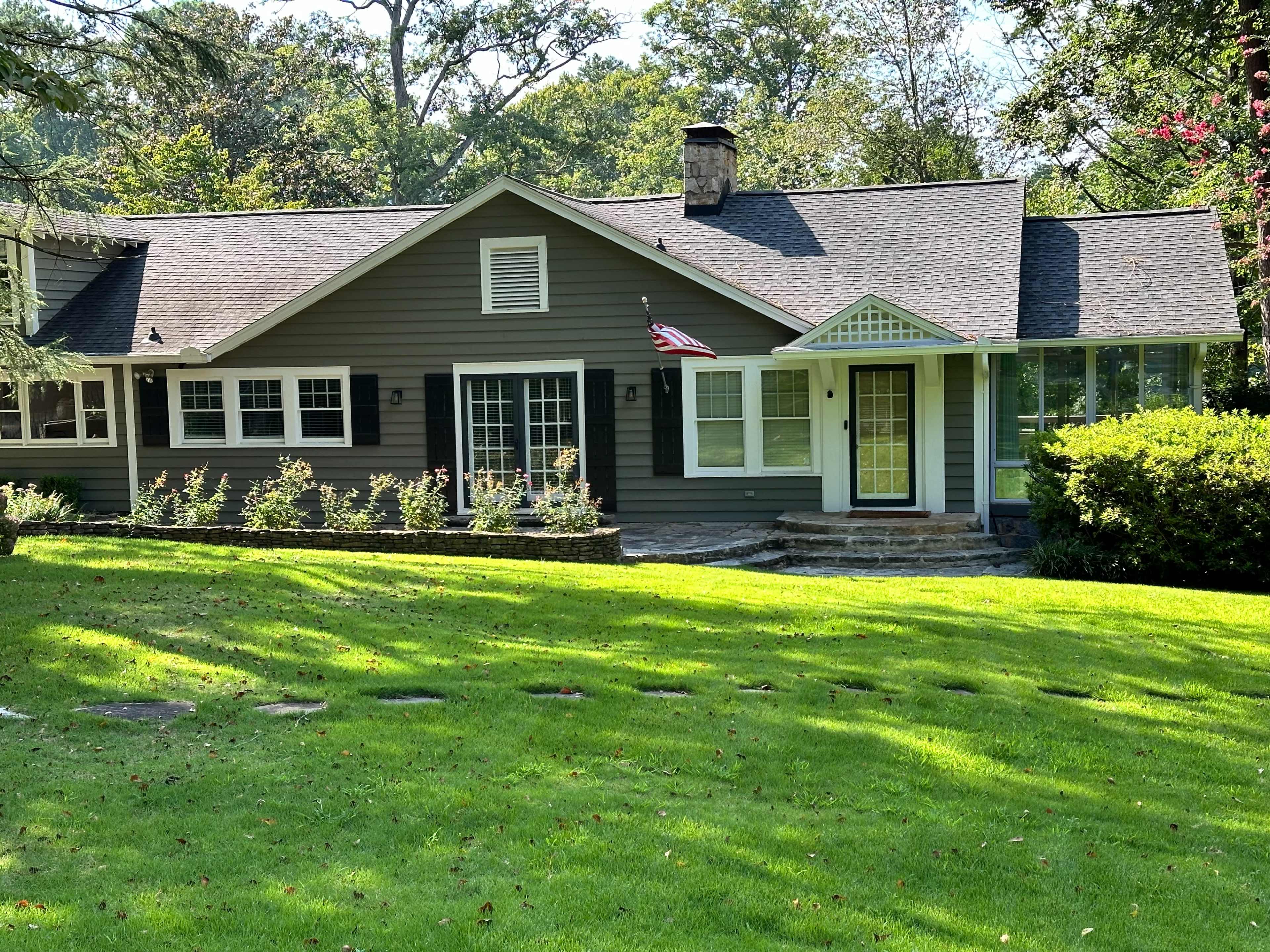 A single-story, gray house with a front porch and an American flag, surrounded by a lawn and trees.