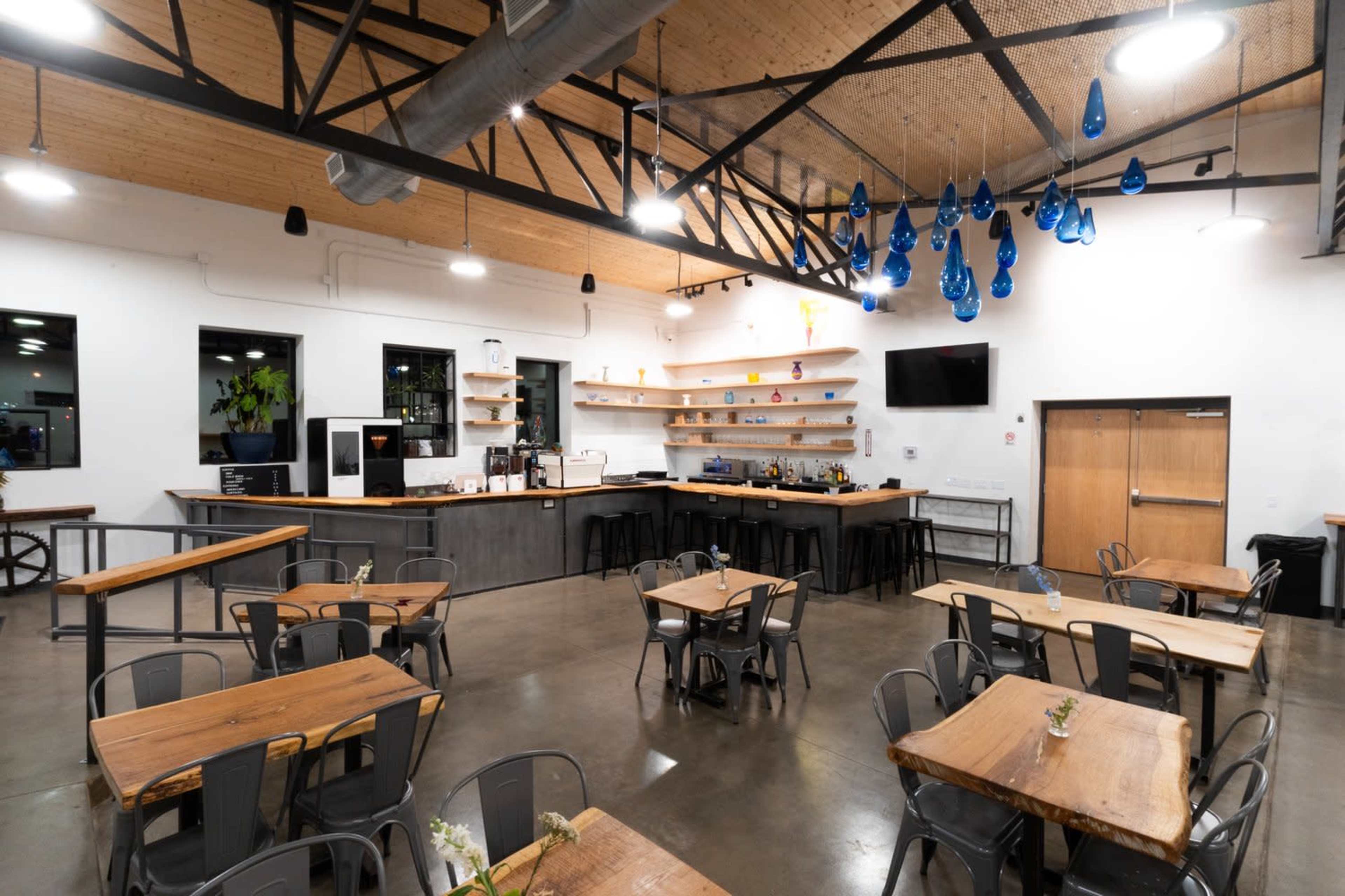 A modern café interior featuring wooden tables, metal chairs, and a bar area with shelves and blue decorative hanging lights.