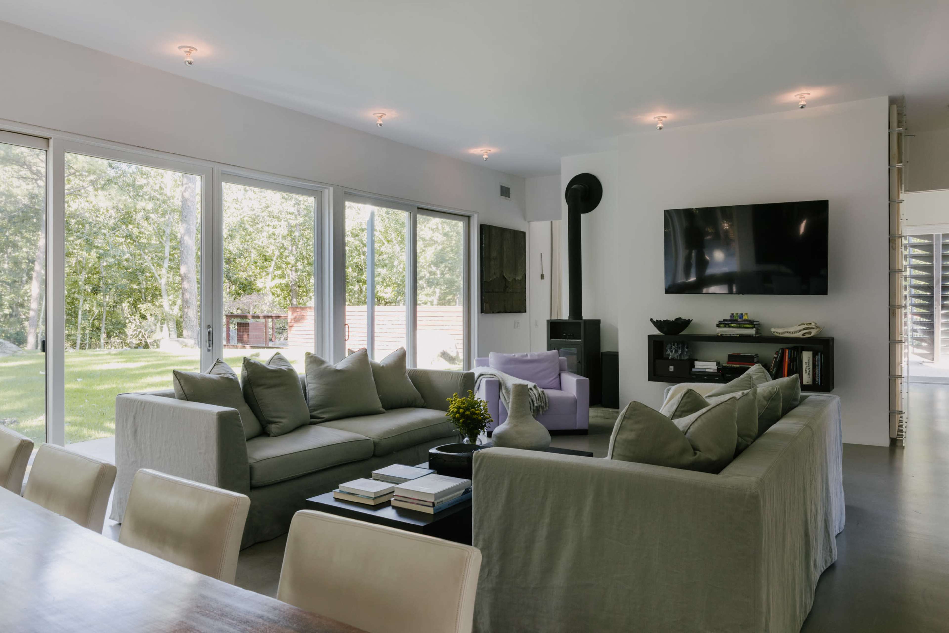 A modern living room features large windows overlooking a grassy area, with a sectional sofa, a purple chair, and a wall-mounted television.