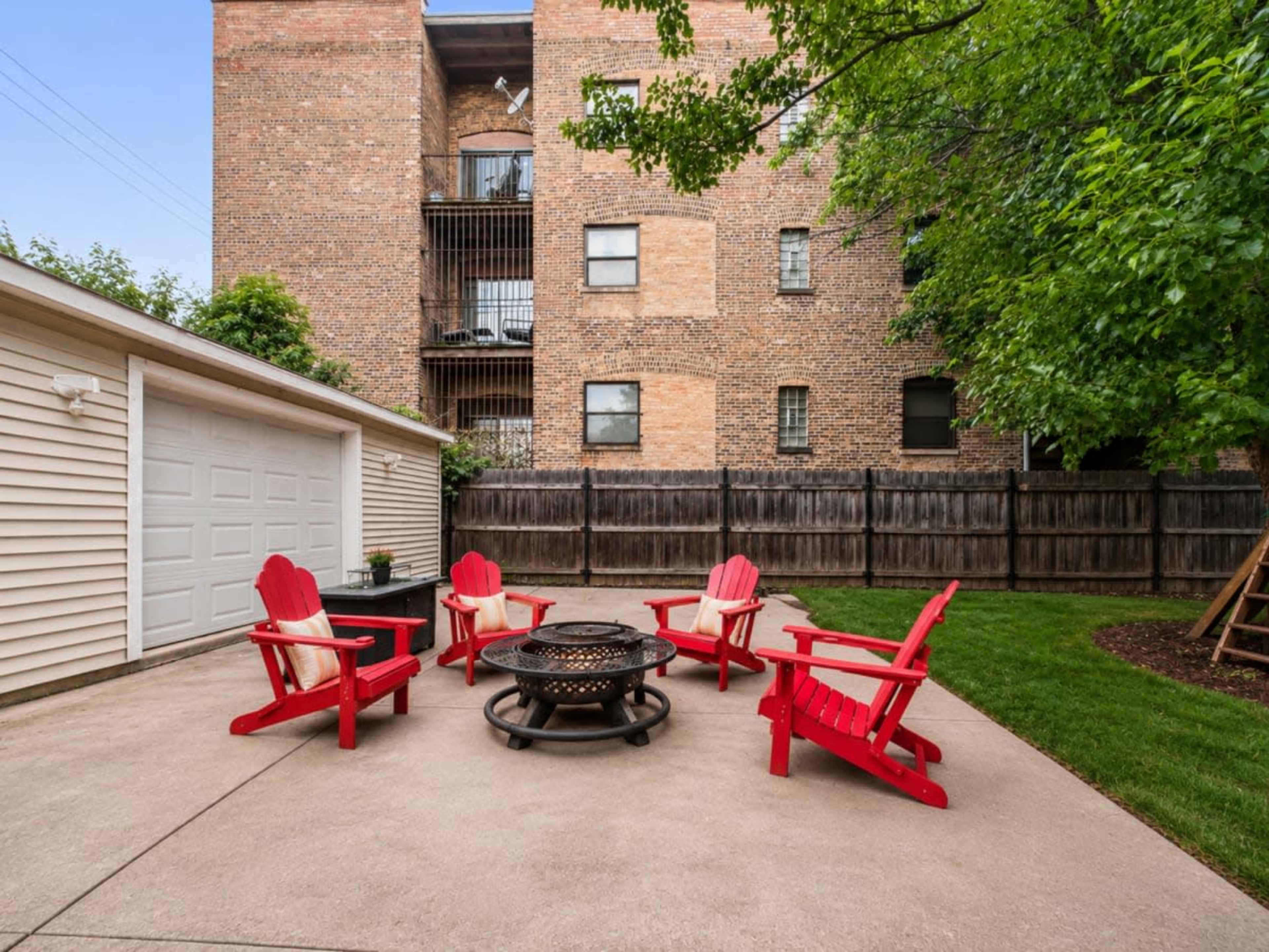 The image shows a concrete patio area with four red Adirondack chairs arranged around a fire pit, bordered by a fence and backed by a brick apartment building.