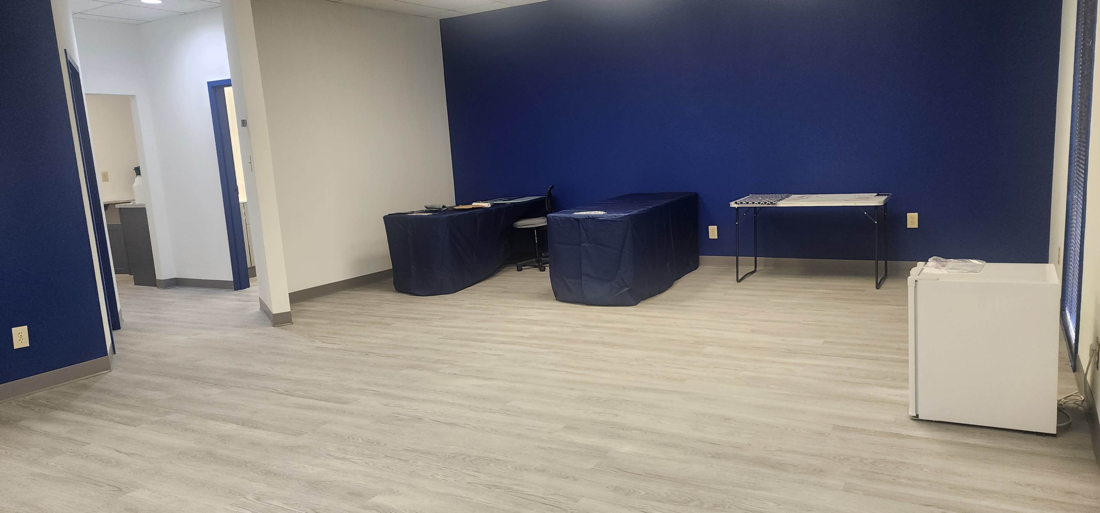 The image shows a minimally furnished room with two tables covered in blue cloth, a chair, and a small white refrigerator against the wall.