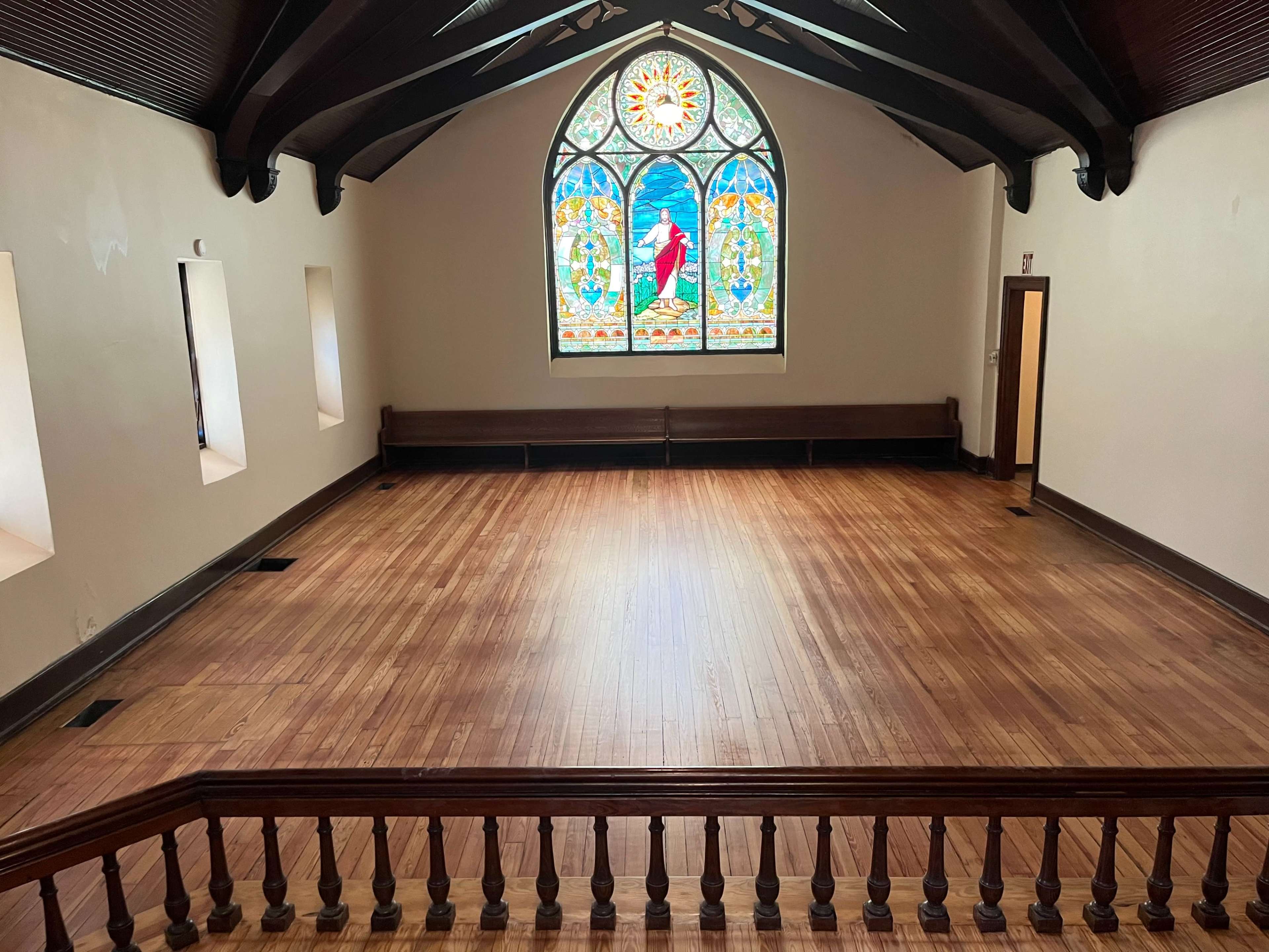The image shows a spacious, empty room with hardwood flooring, featuring a stained glass window at one end and benches along the walls.