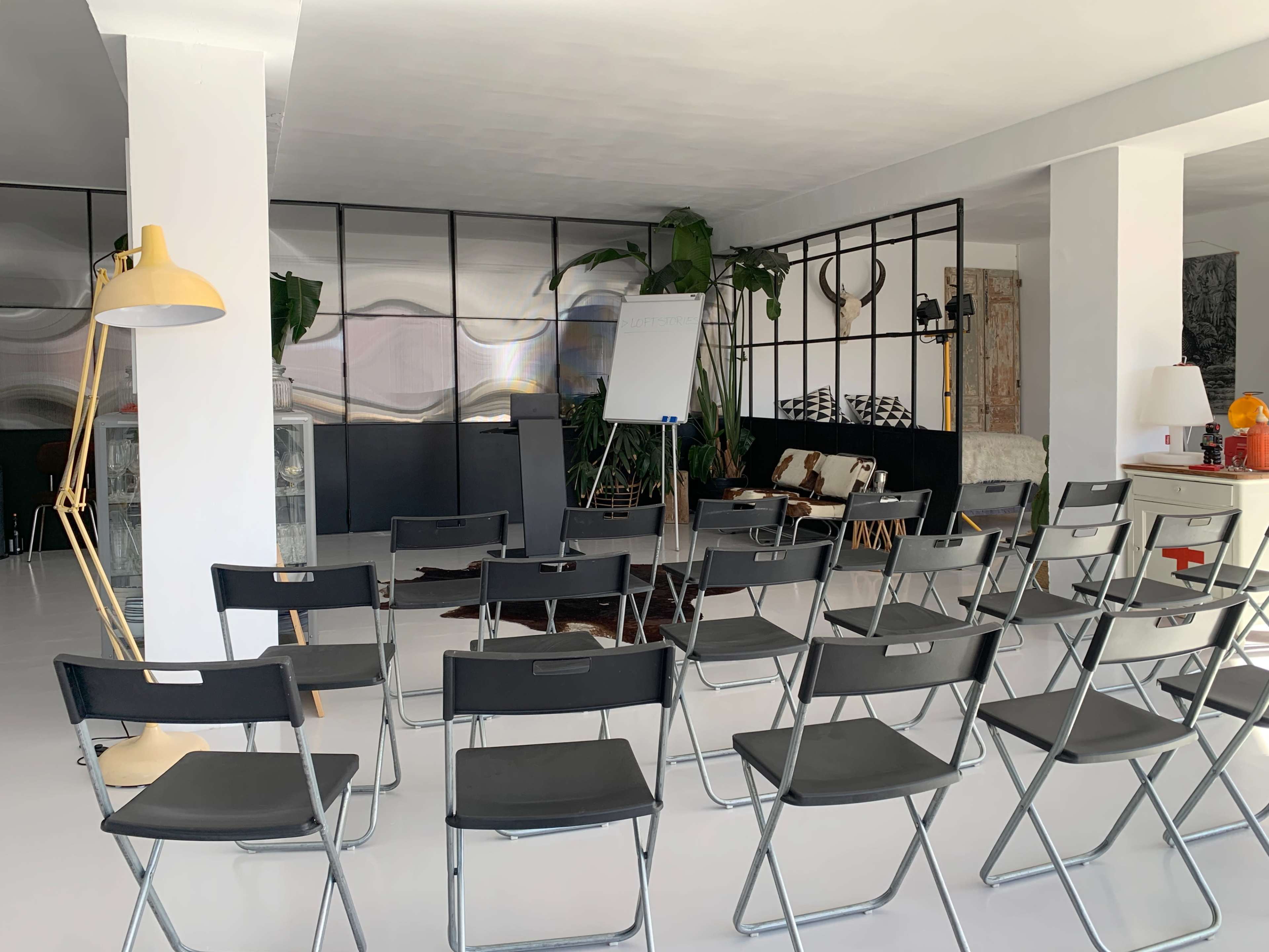 A minimalist meeting room features several black folding chairs arranged in front of a whiteboard, surrounded by plants and various furnishings.