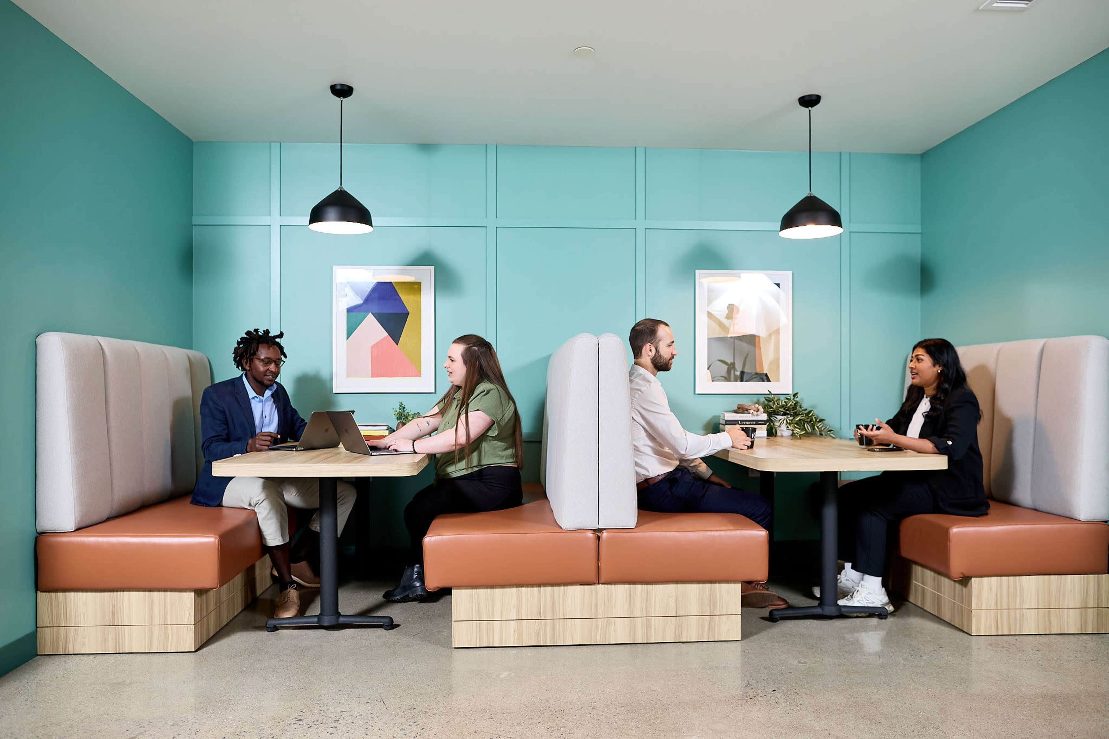 Four people are seated at two separate booths in a modern, brightly colored workspace, engaged in conversation and working on laptops.