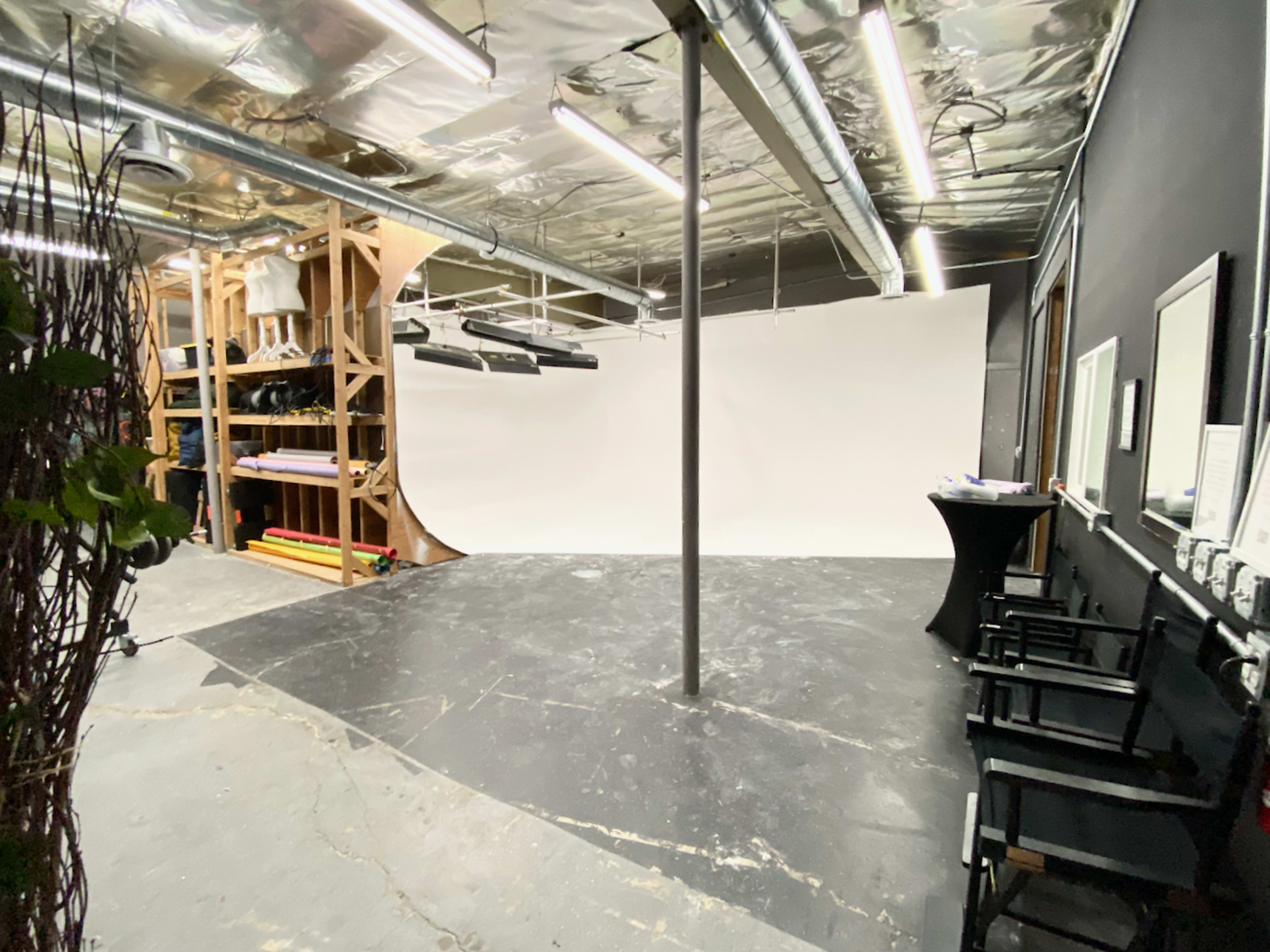 The image shows a photography studio featuring a seamless white backdrop, a wooden shelving unit, and several director's chairs arranged on a black floor.