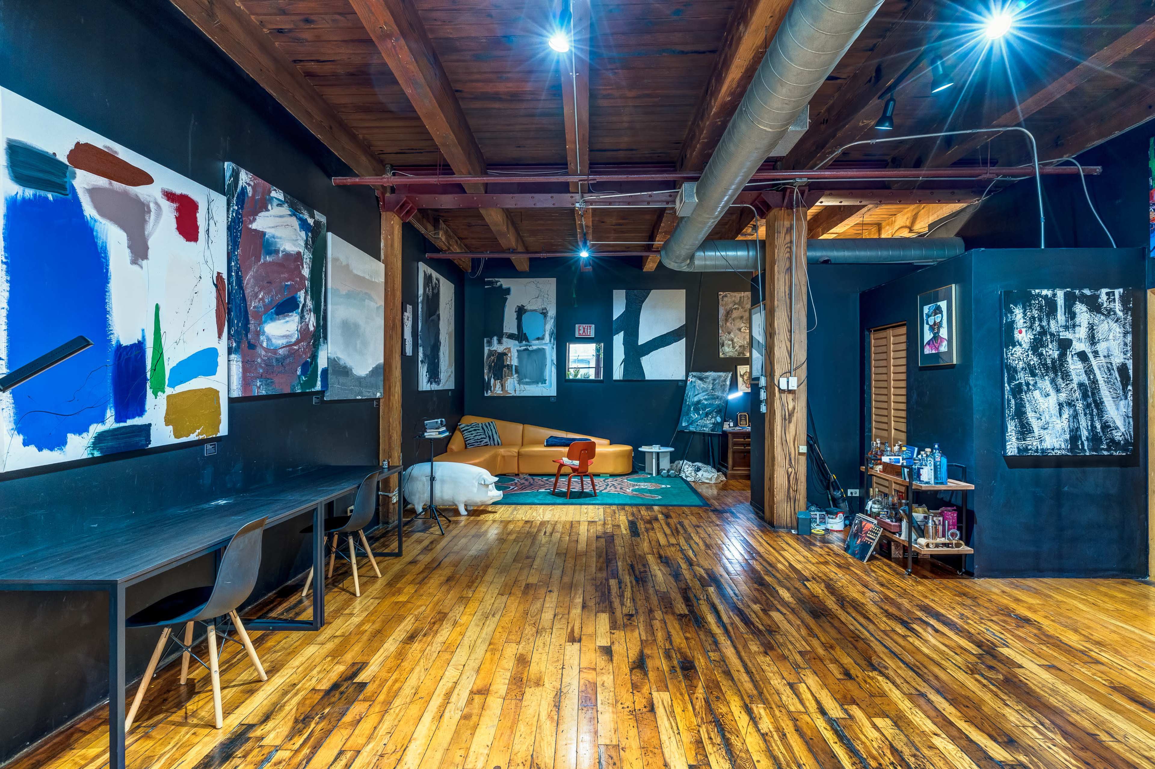 A spacious room with wooden floors, exposed wooden beams, and several abstract art pieces on dark walls, along with a couch and a dining table set against the backdrop.