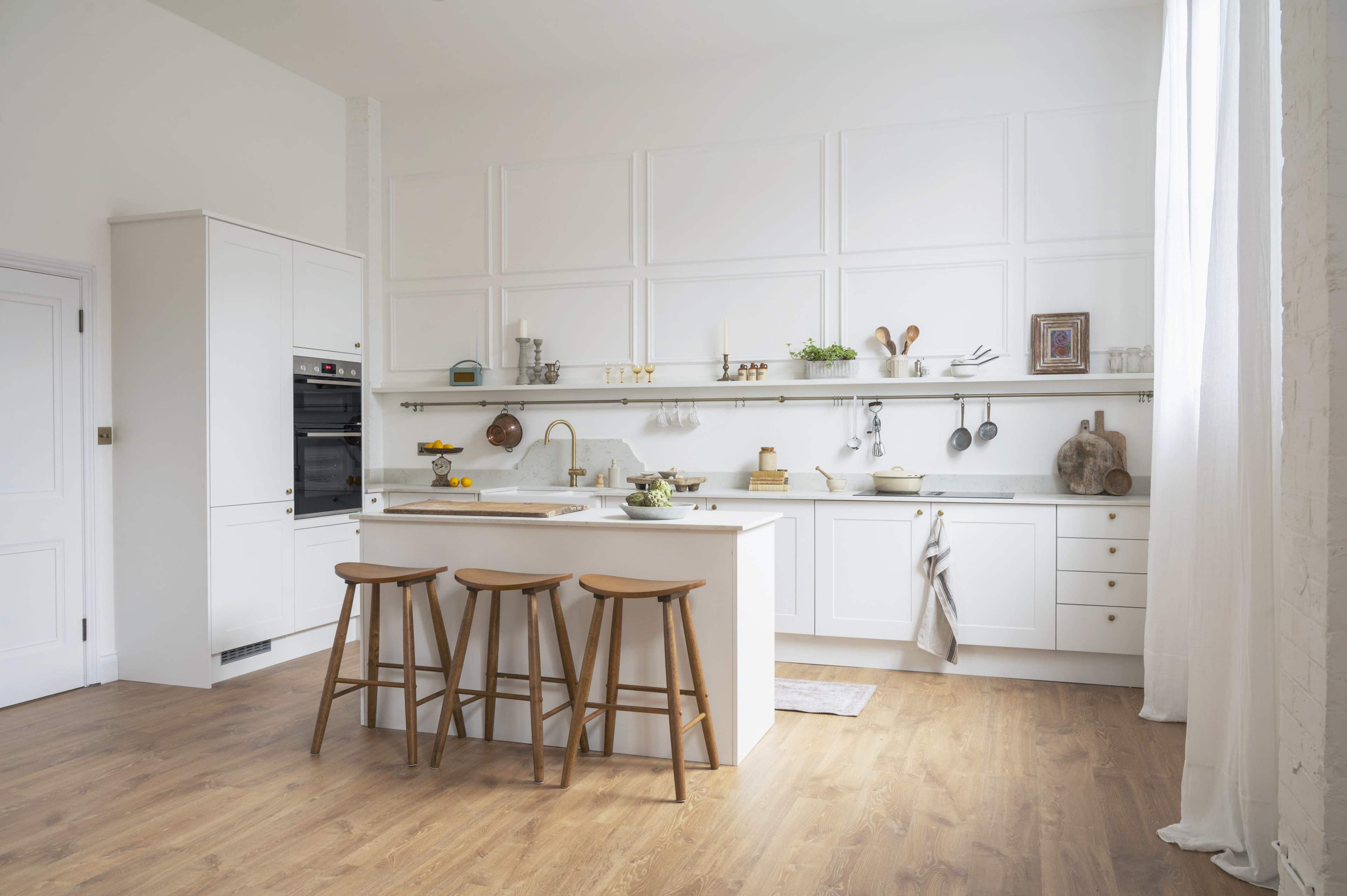 The image shows a modern kitchen featuring white cabinetry, a central island with three wooden stools, and wooden flooring.