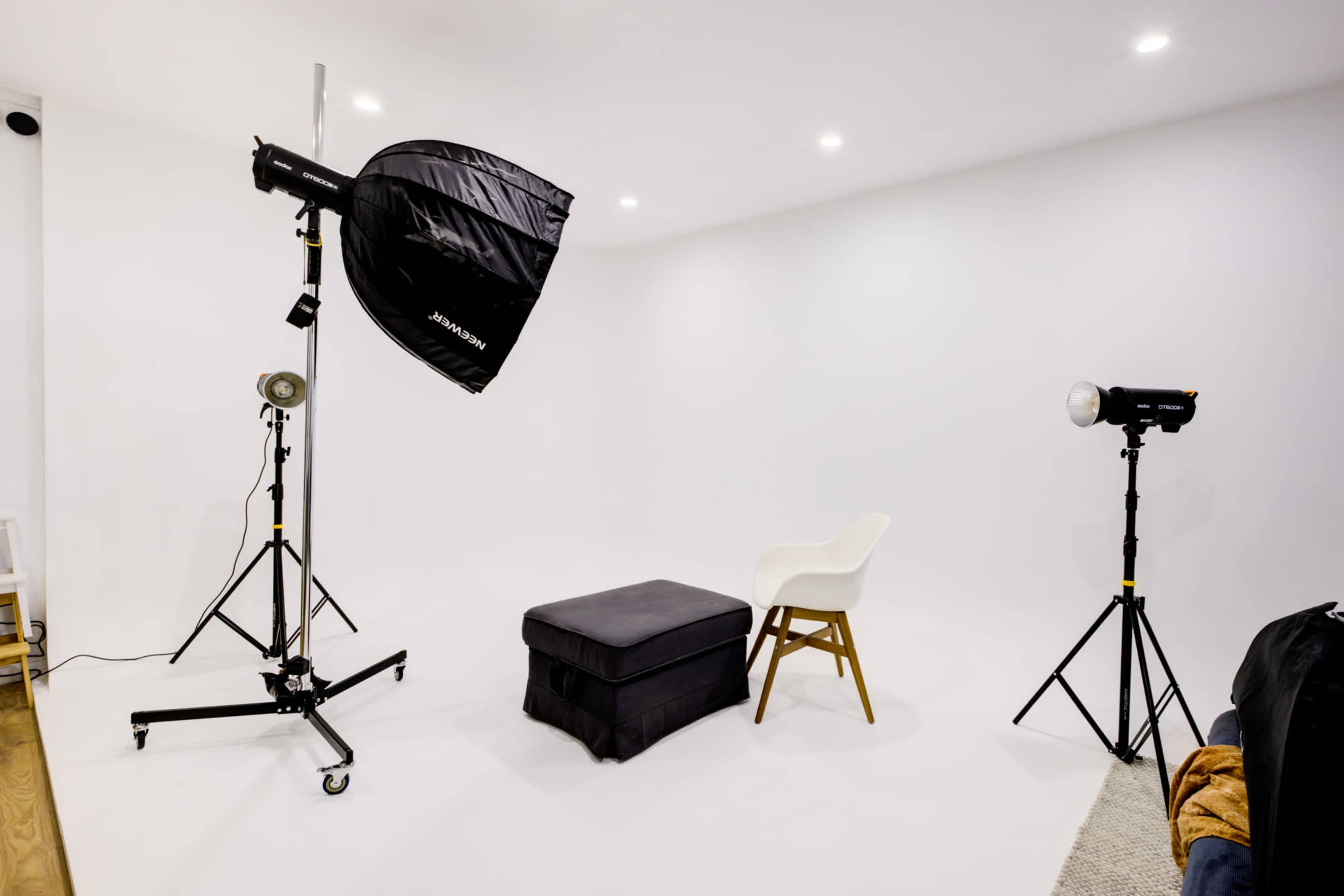 A photography studio with a black softbox light, a white chair, a dark ottoman, and two light stands against a white backdrop.