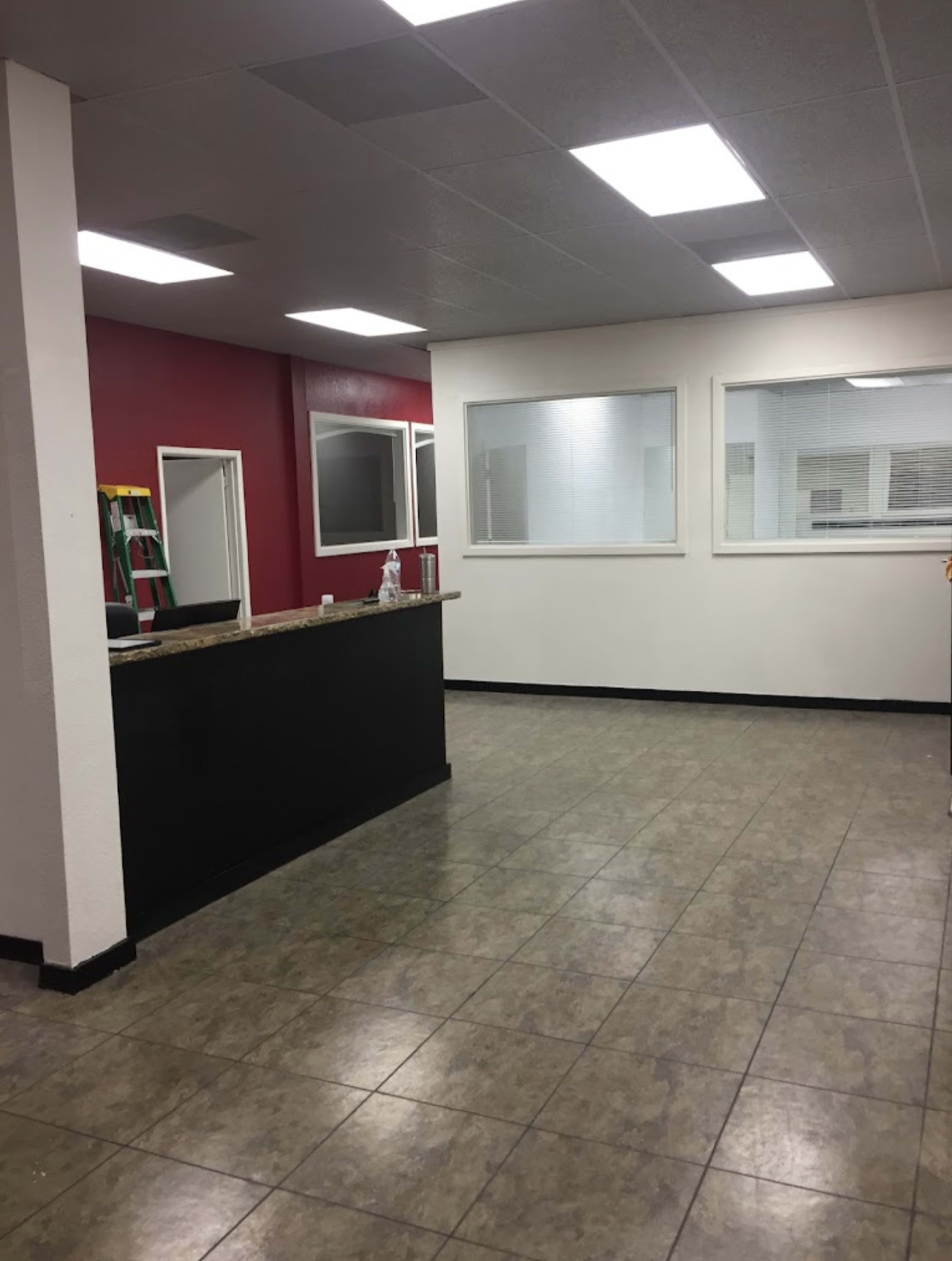 The image shows a partially furnished office space with a front desk, large windows, and a red accent wall.