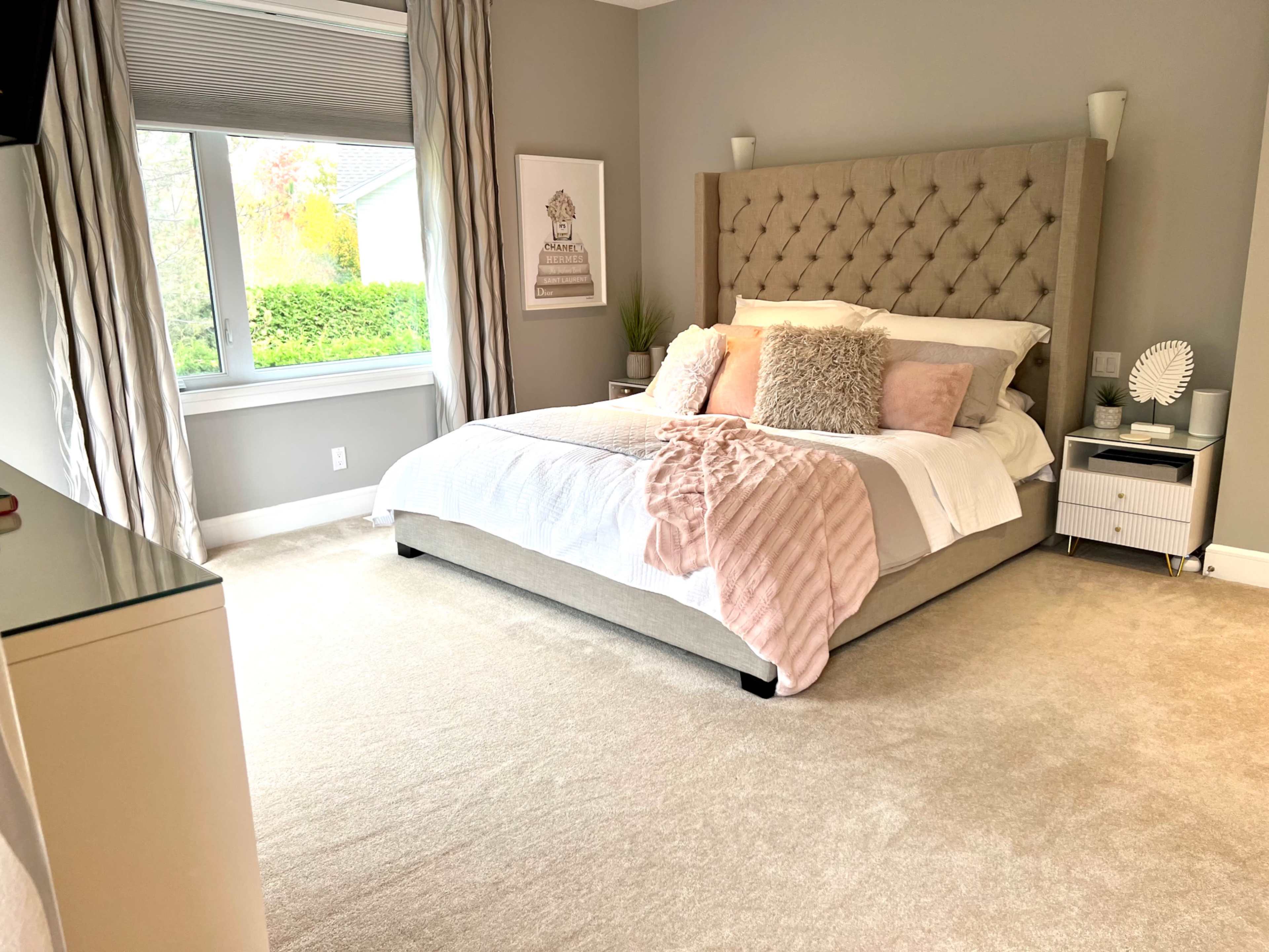 A neatly arranged bedroom features a queen-sized bed with a tufted headboard, soft bedding, and decorative pillows, alongside a window with sheer curtains and a small nightstand.