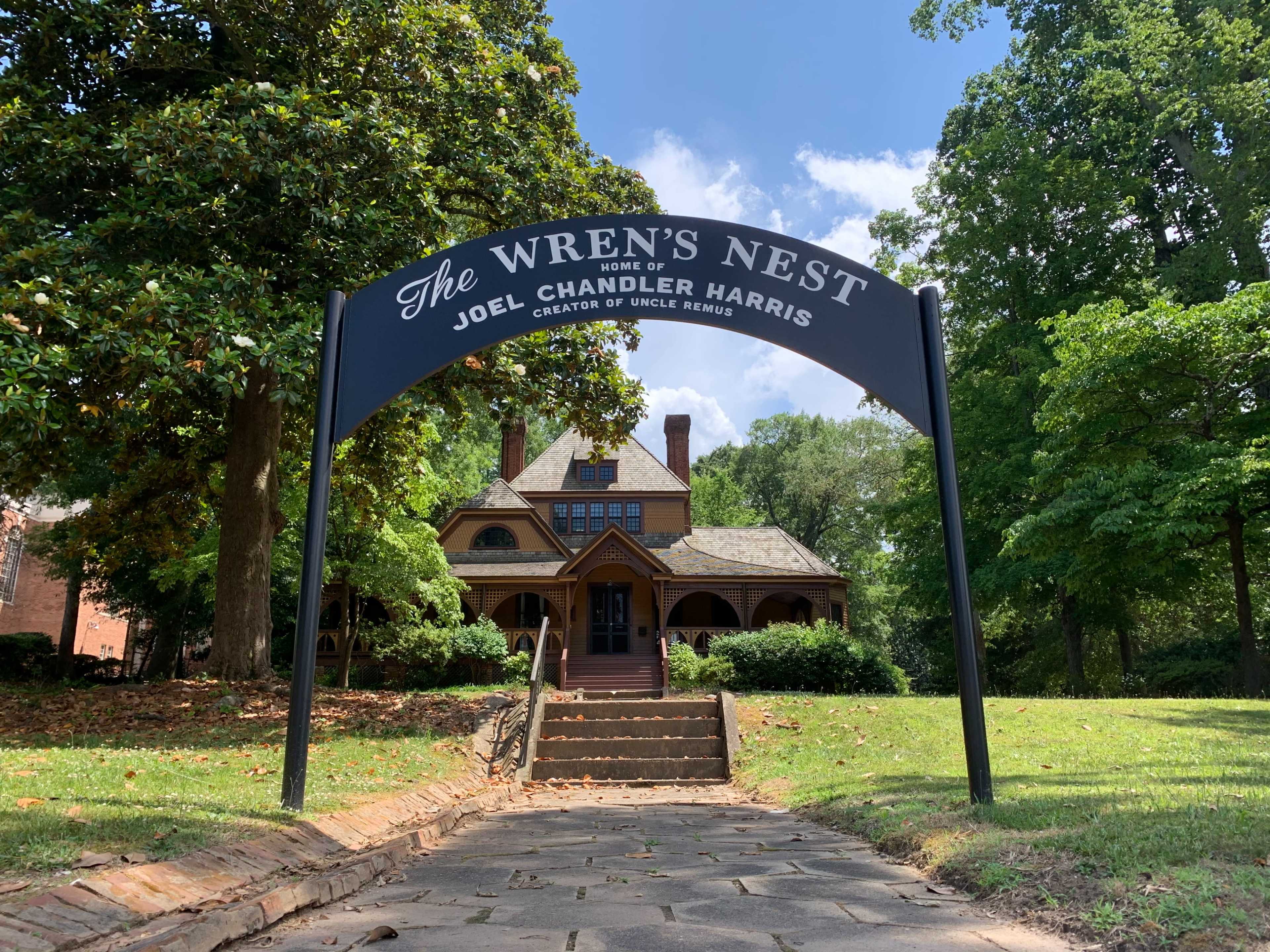 The image shows a historic house with a sign reading "The Wren's Nest, home of Joel Chandler Harris, creator of Uncle Remus," surrounded by green trees and a clear blue sky.