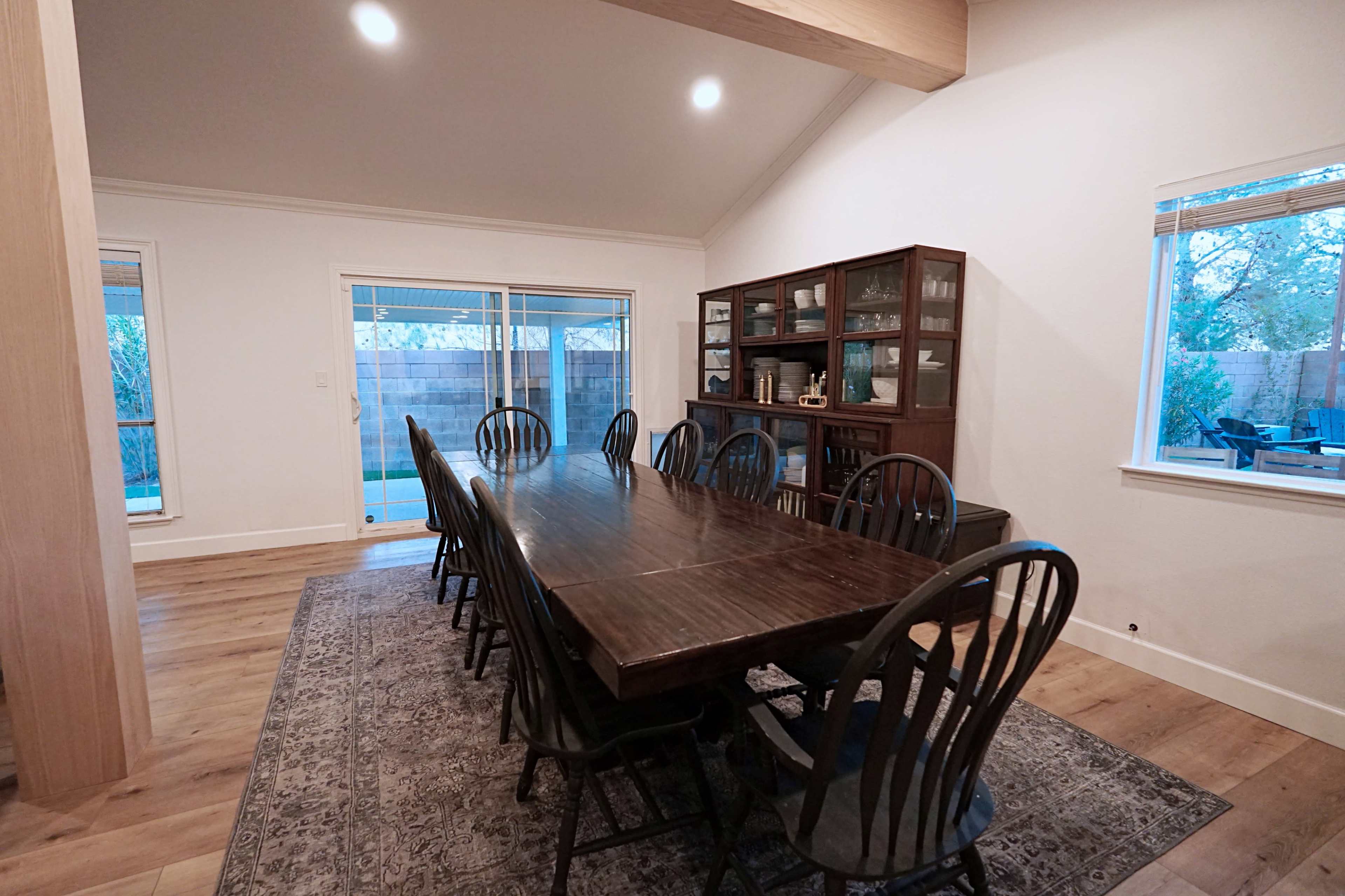 A spacious dining room featuring a long wooden table surrounded by black chairs, with a glass-fronted cabinet and large windows that provide natural light.