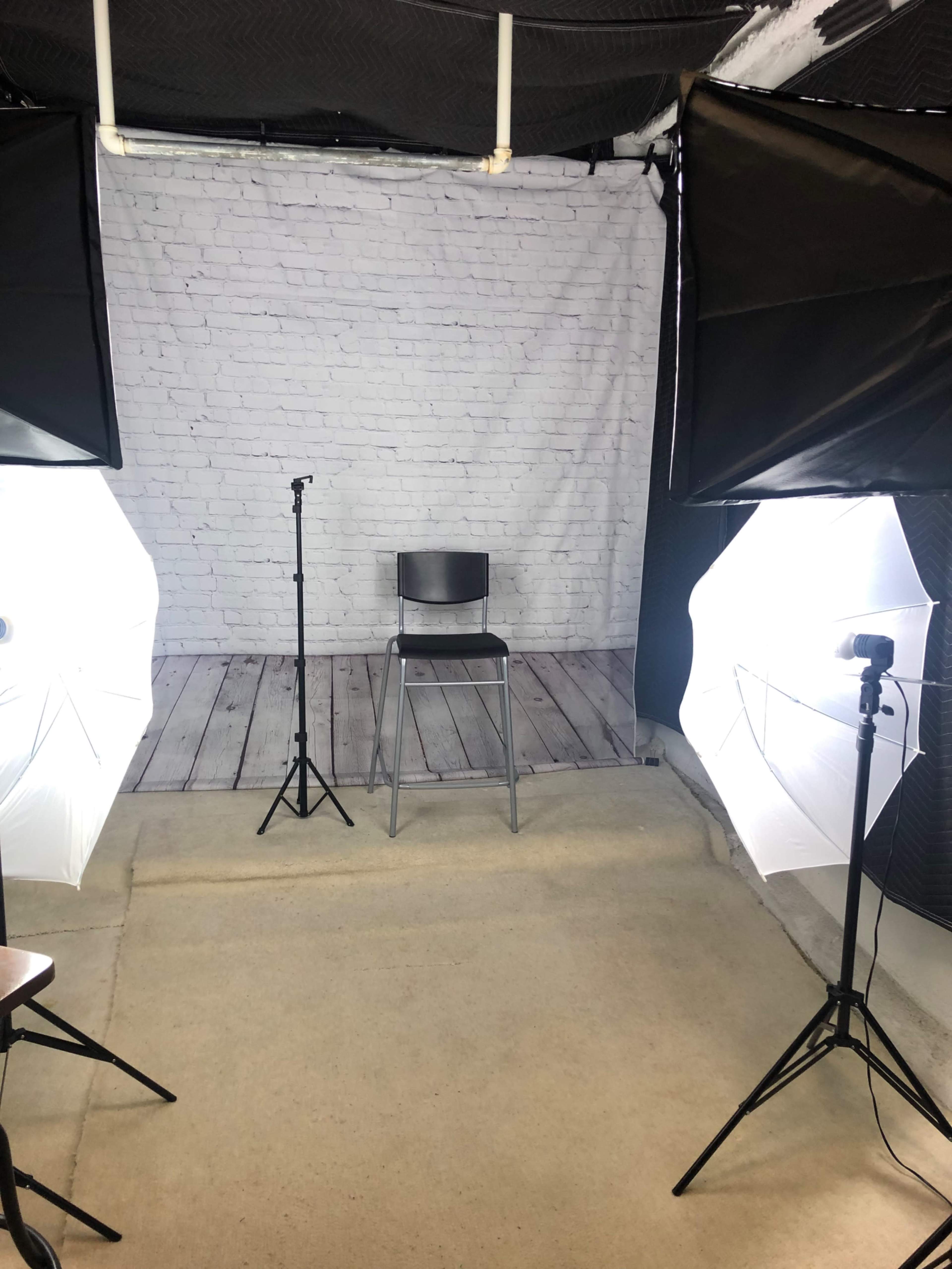 A photography setup featuring a single empty chair in front of a white brick wall backdrop, illuminated by two softbox lights on either side.