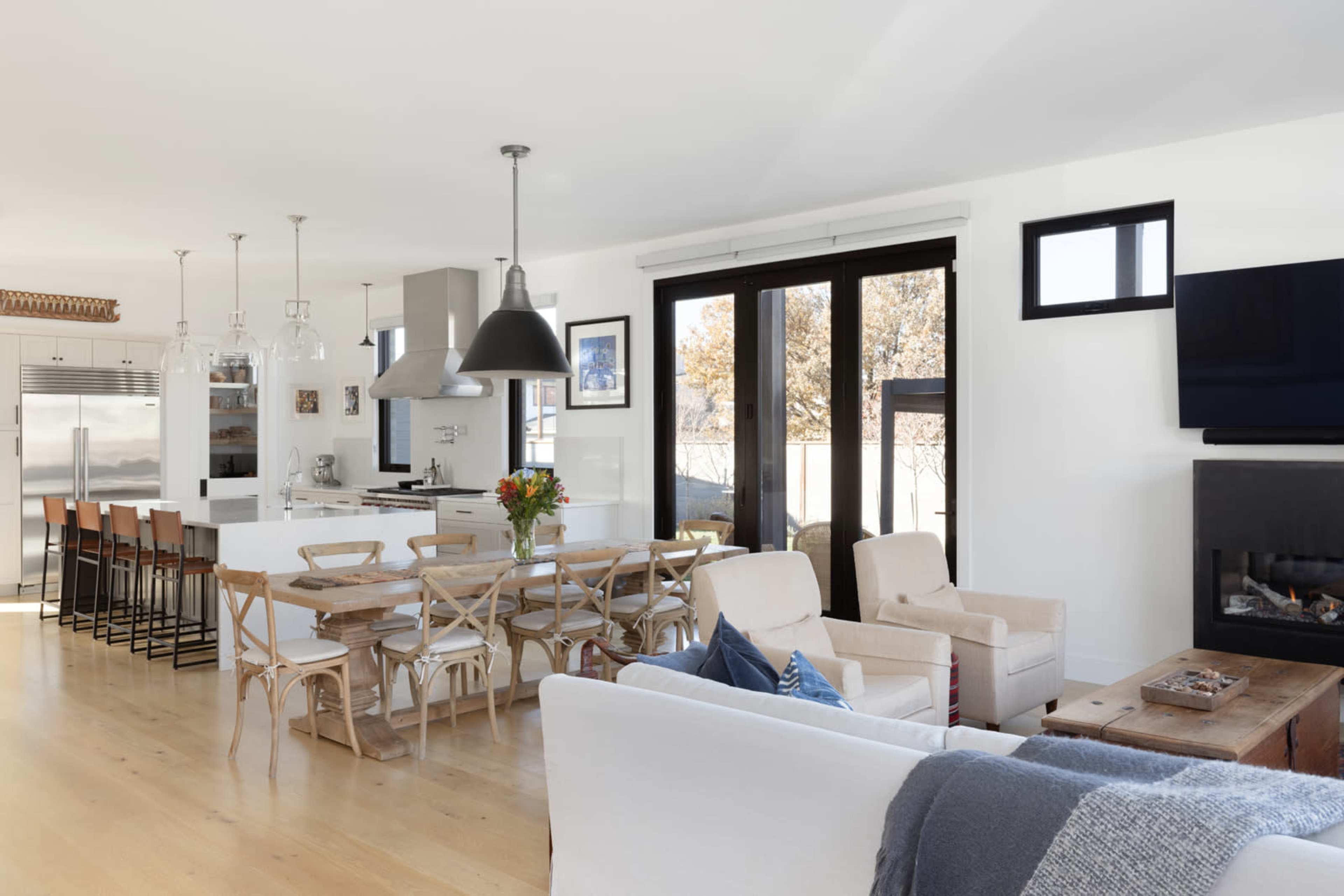 The image shows a modern, open-concept living area with a kitchen, dining table, and seating area featuring large windows and light-colored furnishings.