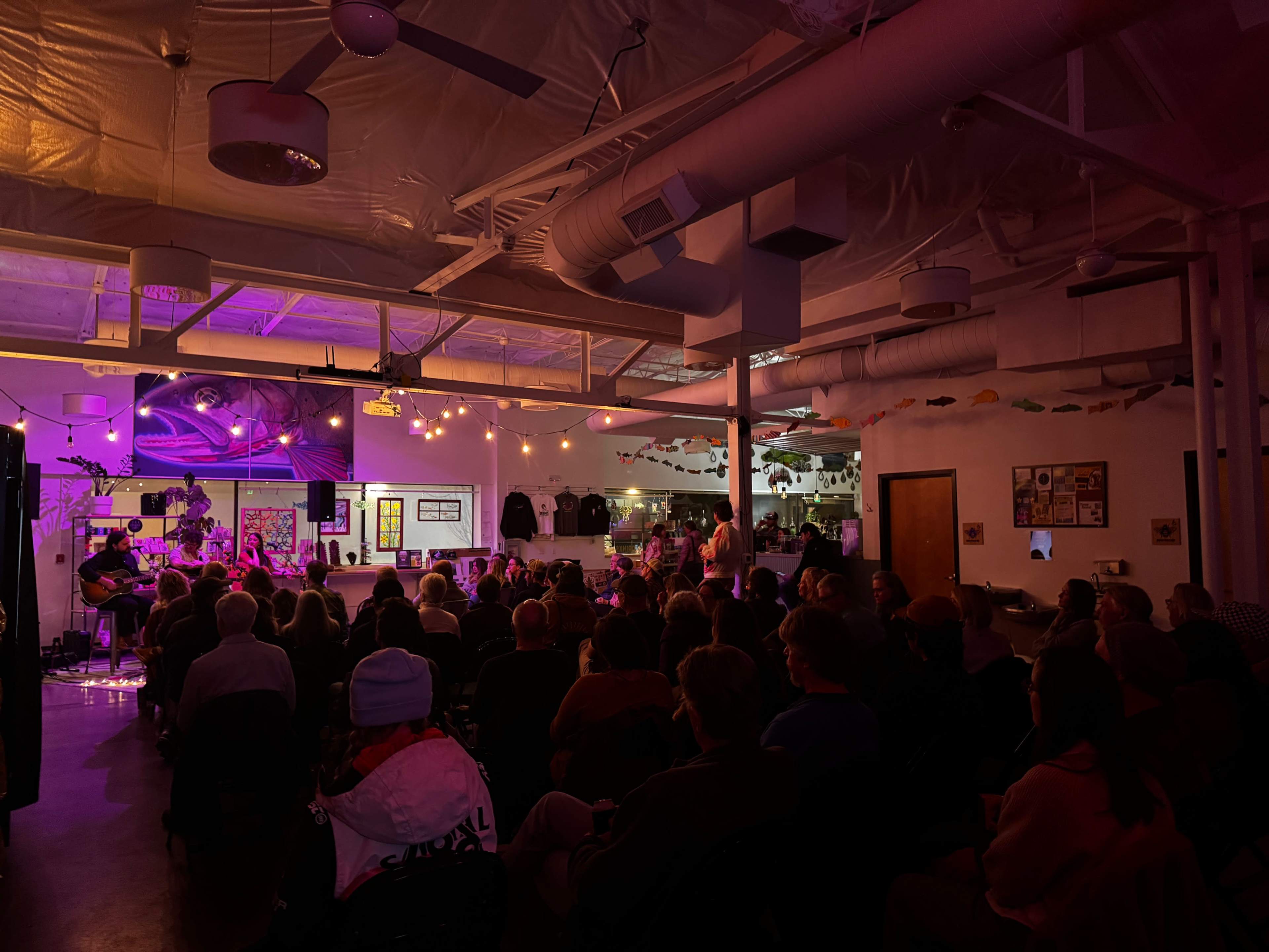 A crowd sits in a dimly lit venue, watching a live music performance illuminated by soft purple and warm lights.