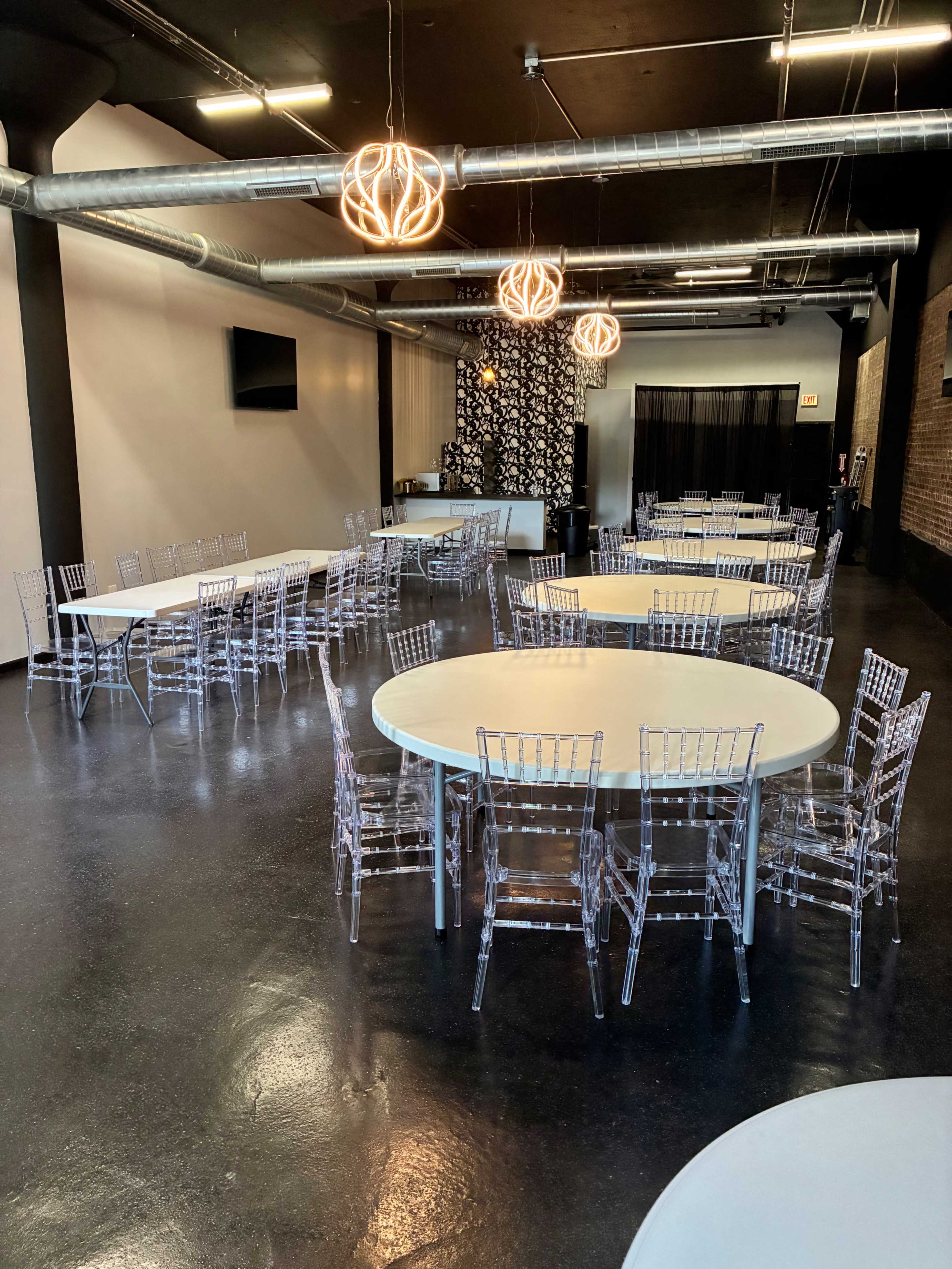 The image shows a spacious event venue featuring several round tables surrounded by clear acrylic chairs, with modern lighting fixtures hanging from the ceiling.