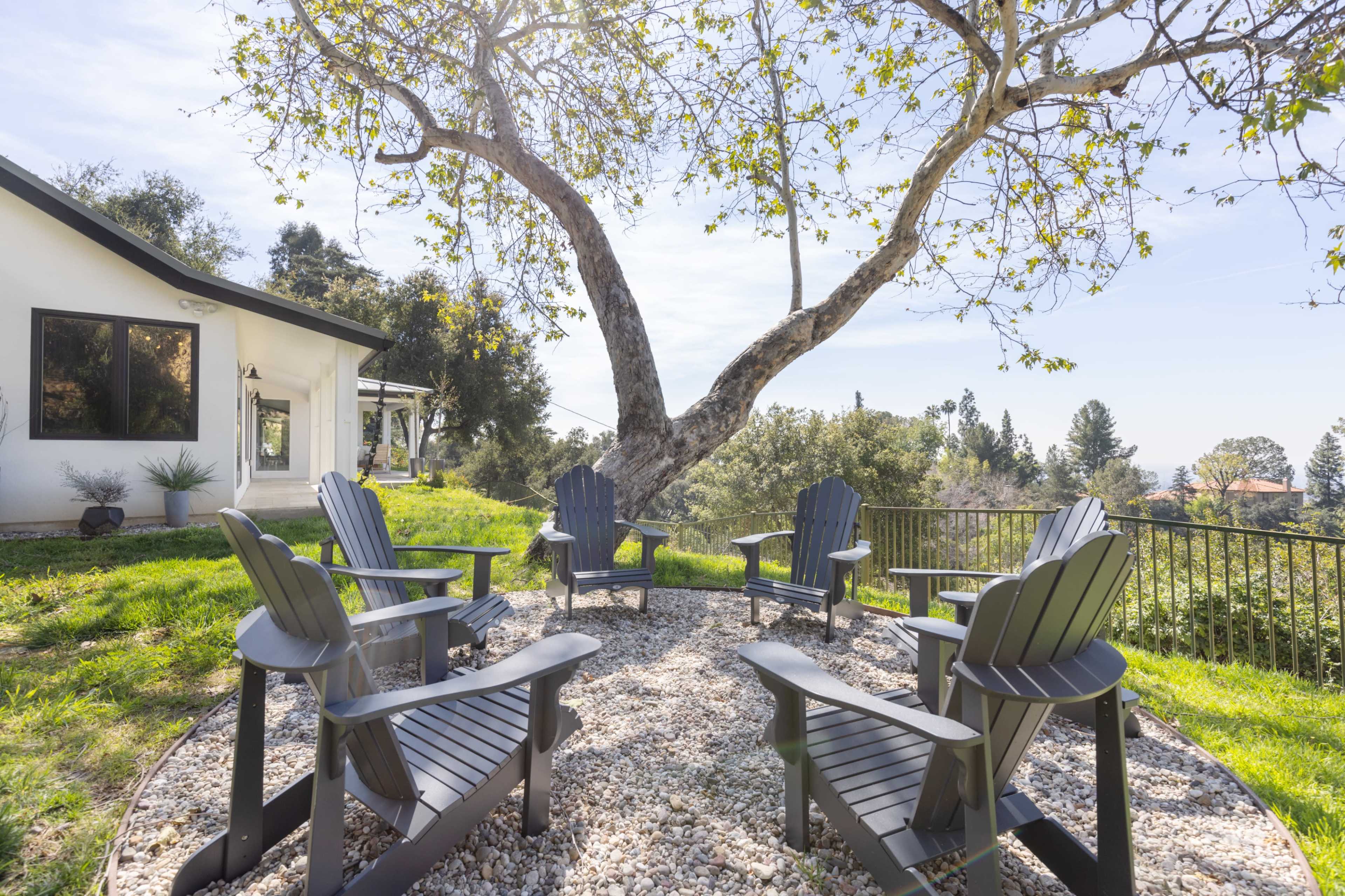 A circular seating area with black Adirondack chairs is surrounded by gravel and overlooks a green landscape and a large tree.