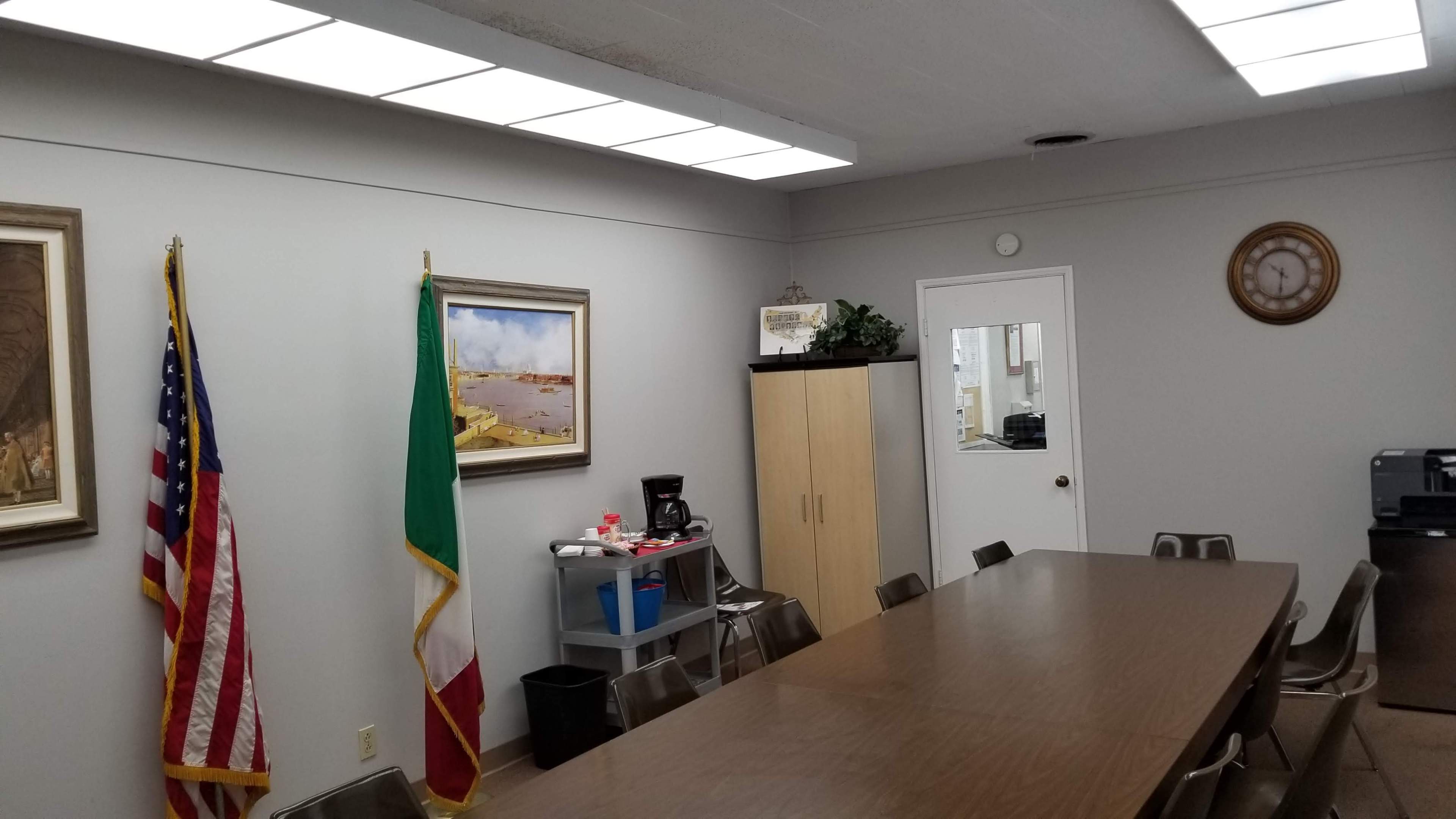 A conference room features a long table surrounded by chairs, with an American flag, an Italian flag, and various wall decor, including artwork and a clock.