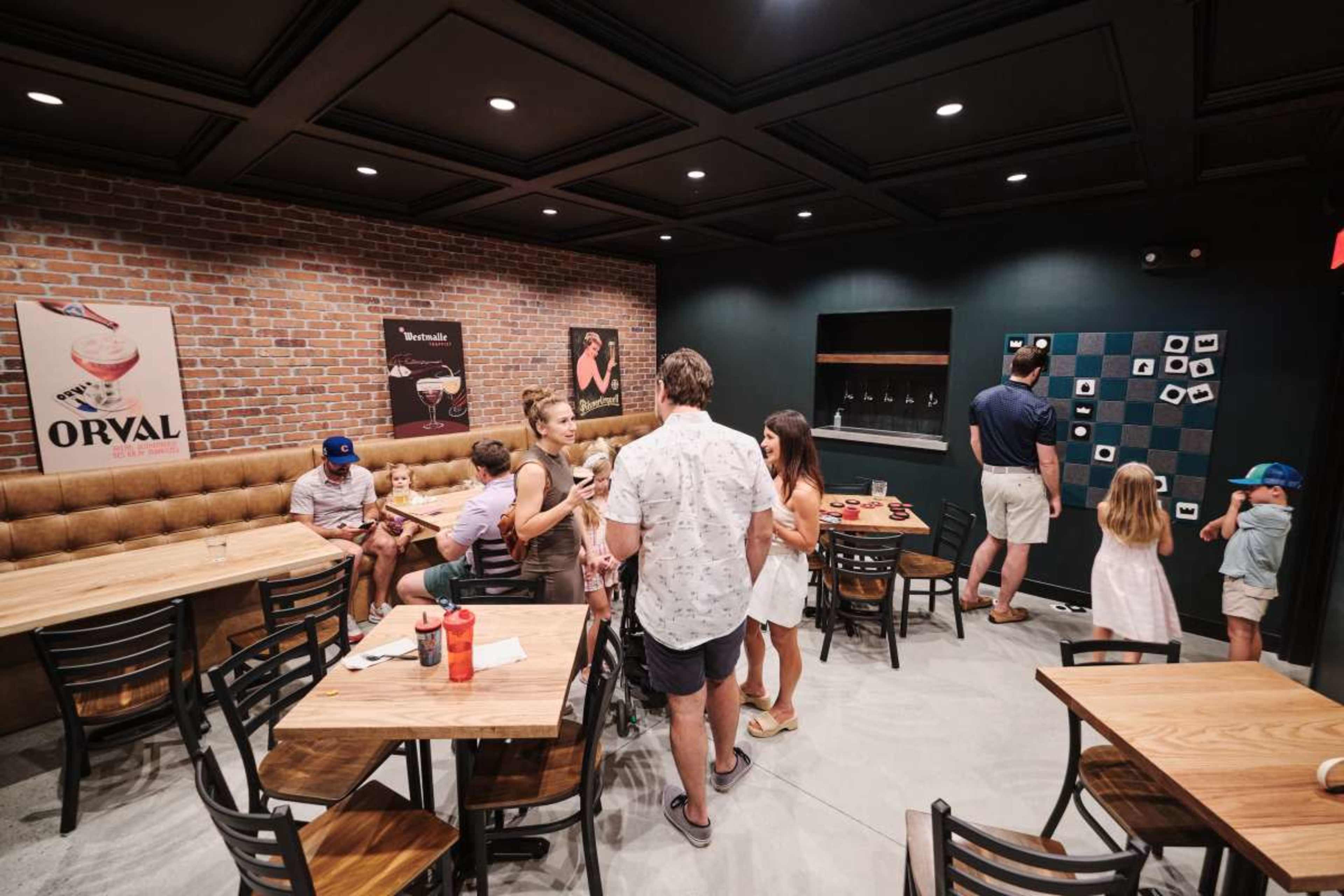 A group of people mingles in a cozy restaurant with brick walls, wooden tables, and a game wall featuring various cards.