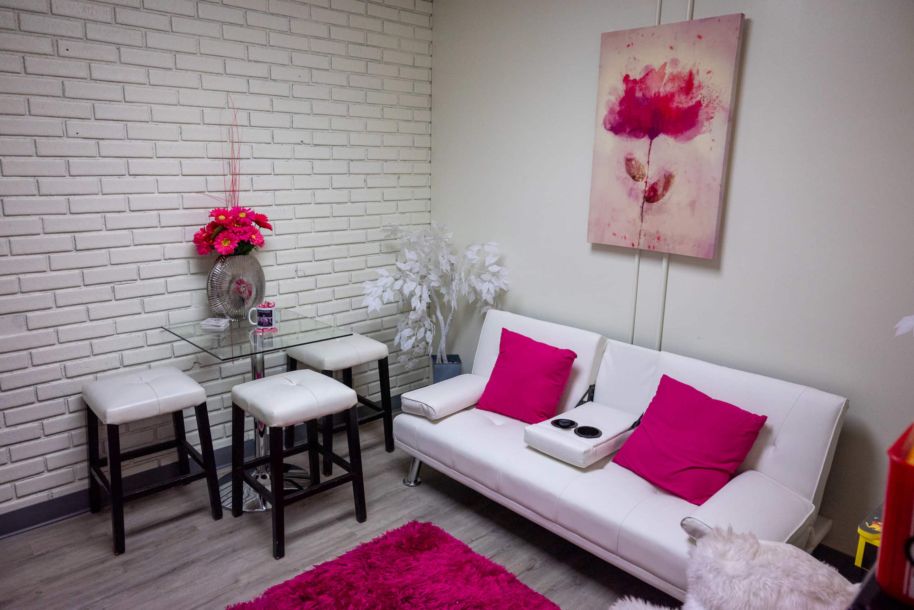 A small lounge area featuring a white sofa with pink cushions, a glass table with two black stools, and a flower-themed painting on the wall.