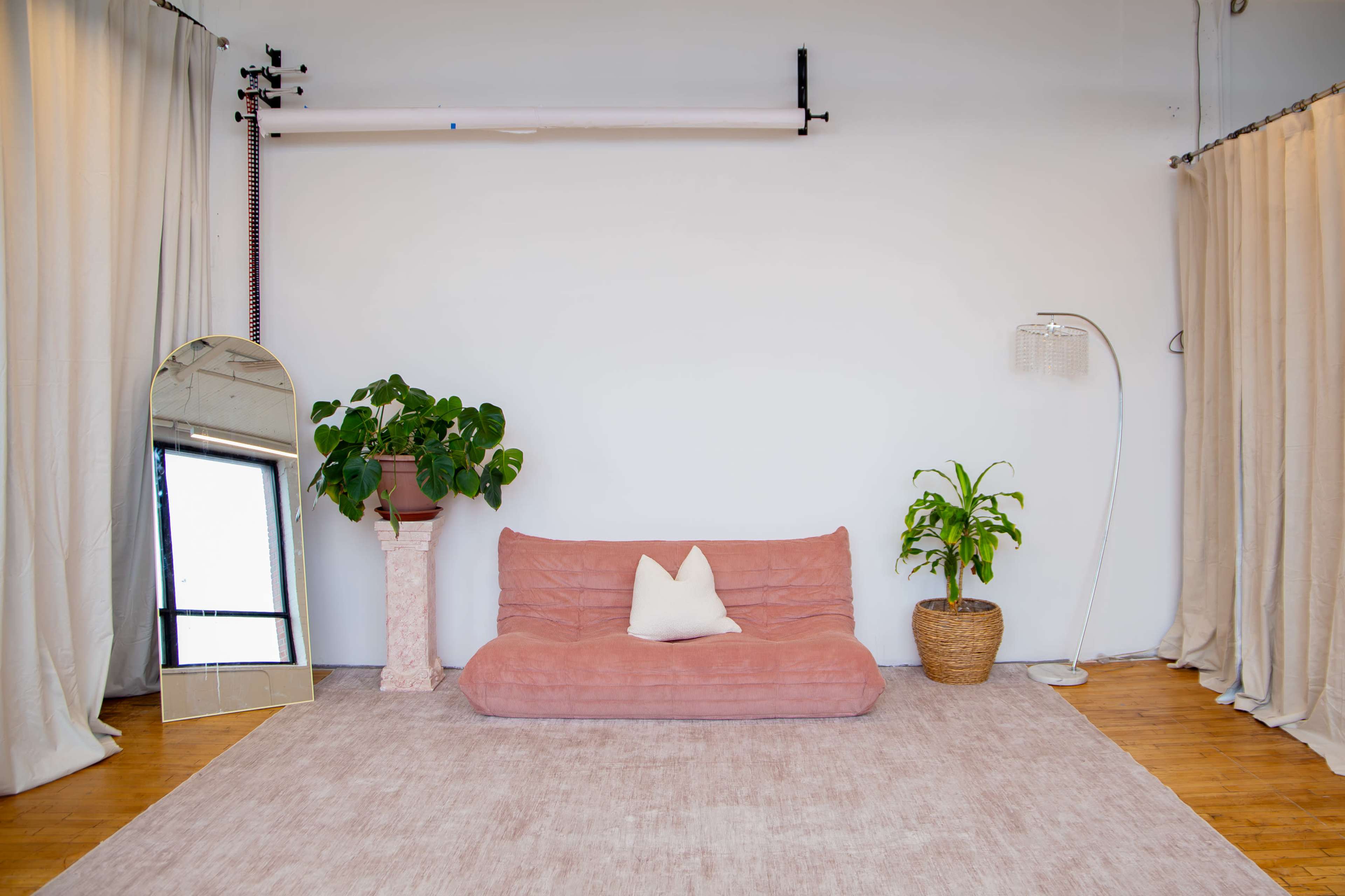 The image shows a minimalist interior featuring a pink sofa, a large mirror, two potted plants, and soft lighting with curtains.