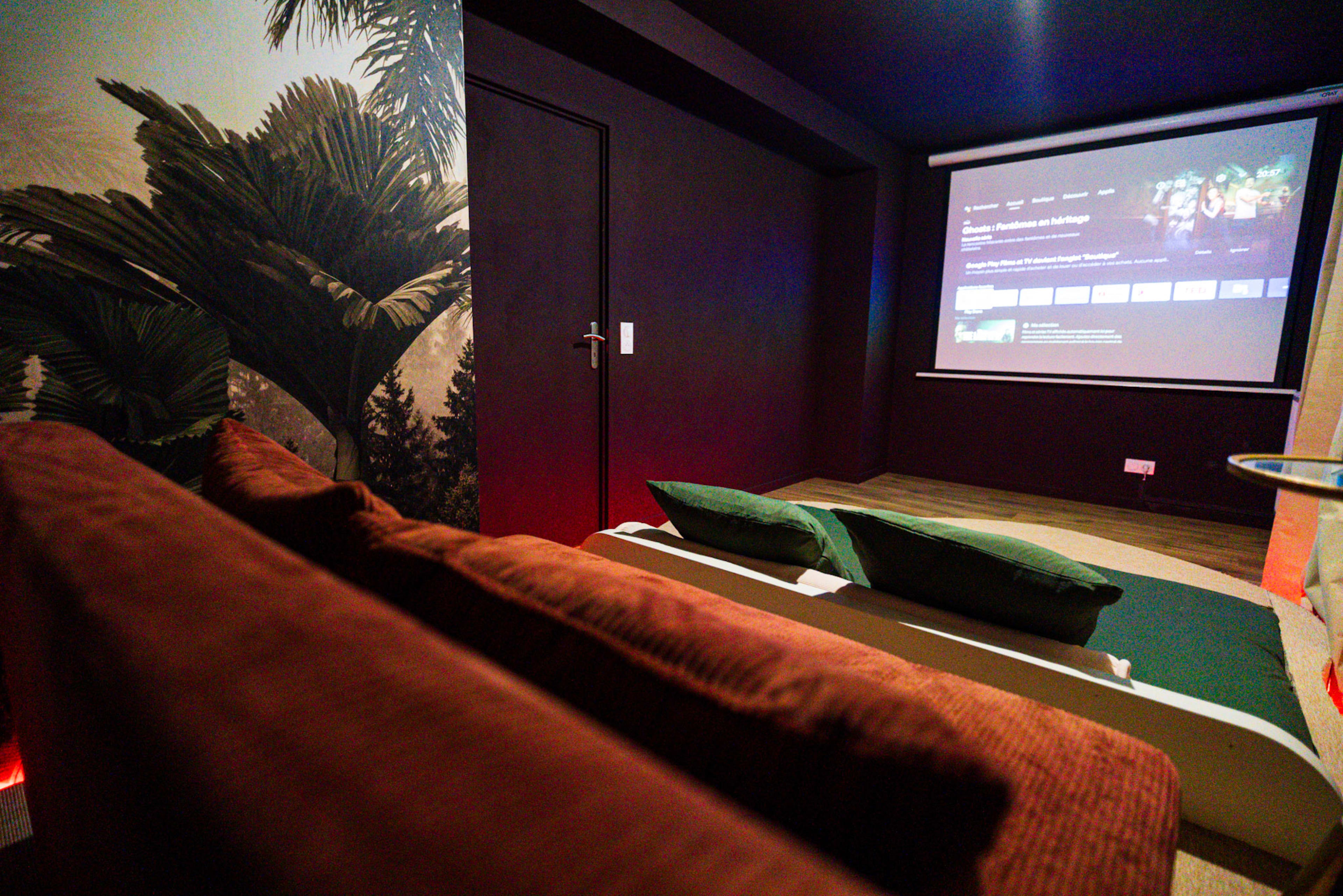 The image shows a cozy home theater setup with a projection screen displaying a menu, plush seating, and dark walls decorated with a tropical mural.