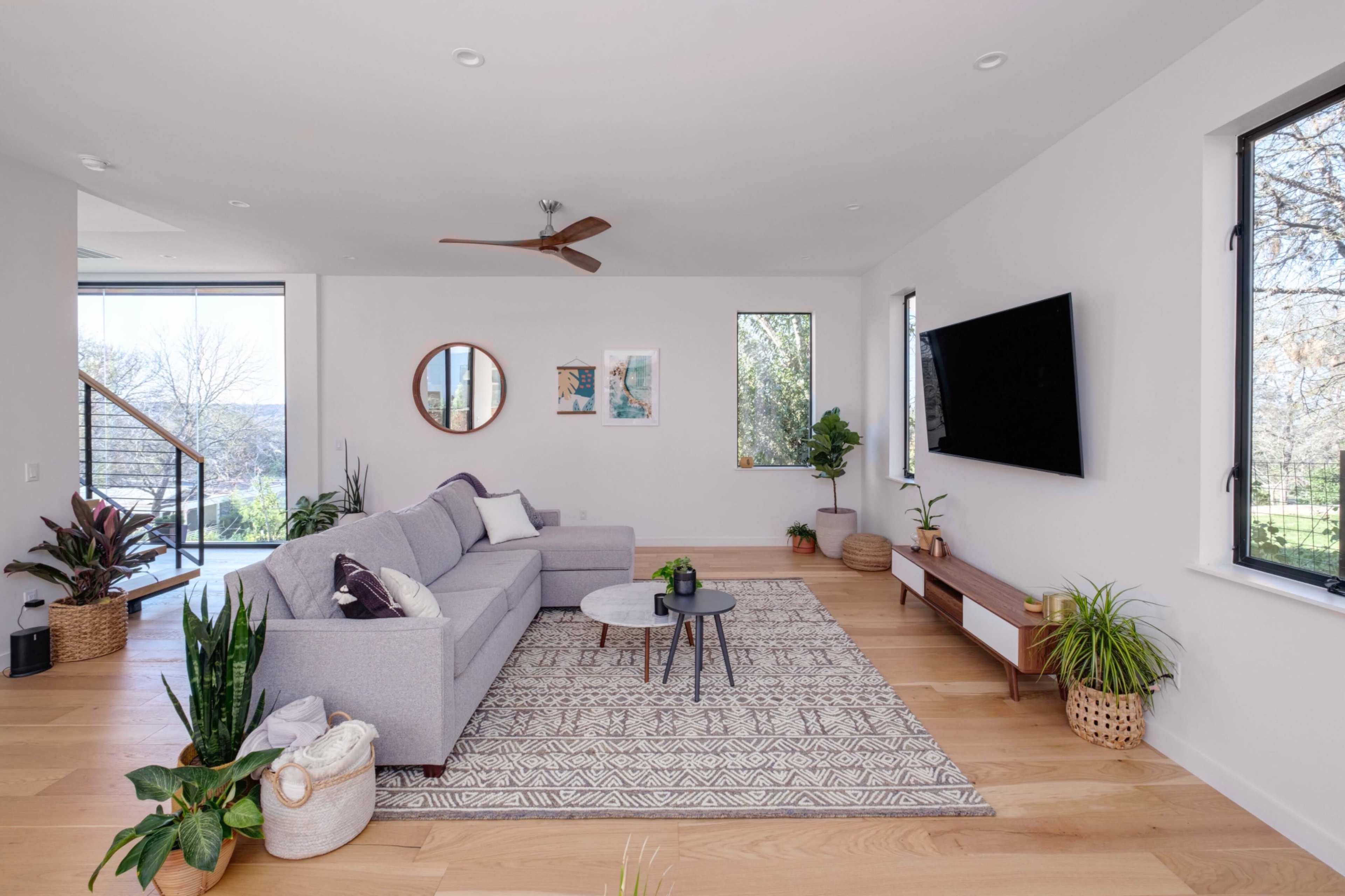 A modern living room features a gray sectional sofa, a round coffee table, and various potted plants, with large windows allowing natural light to enter.