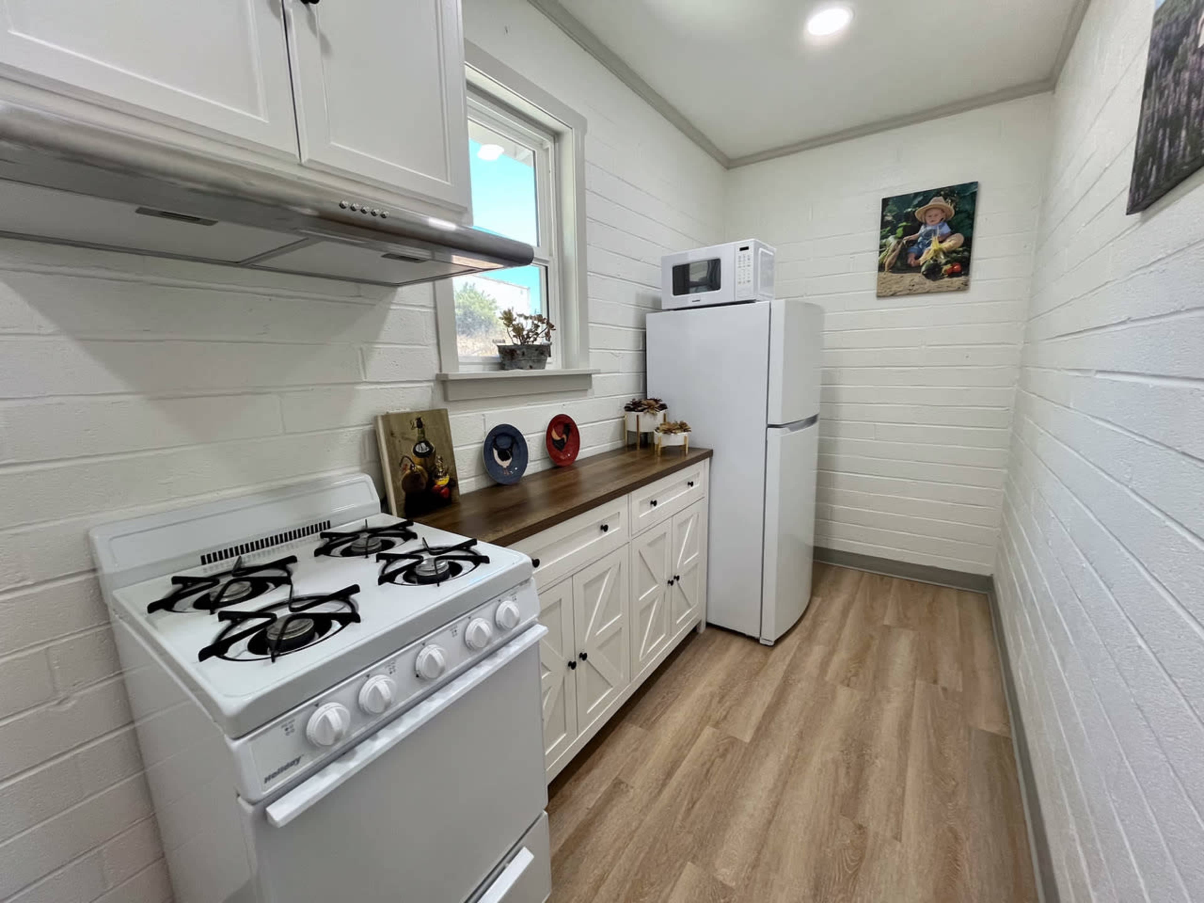 The image shows a small kitchen with white brick walls, a stove, a refrigerator, and a countertop with decorative items.