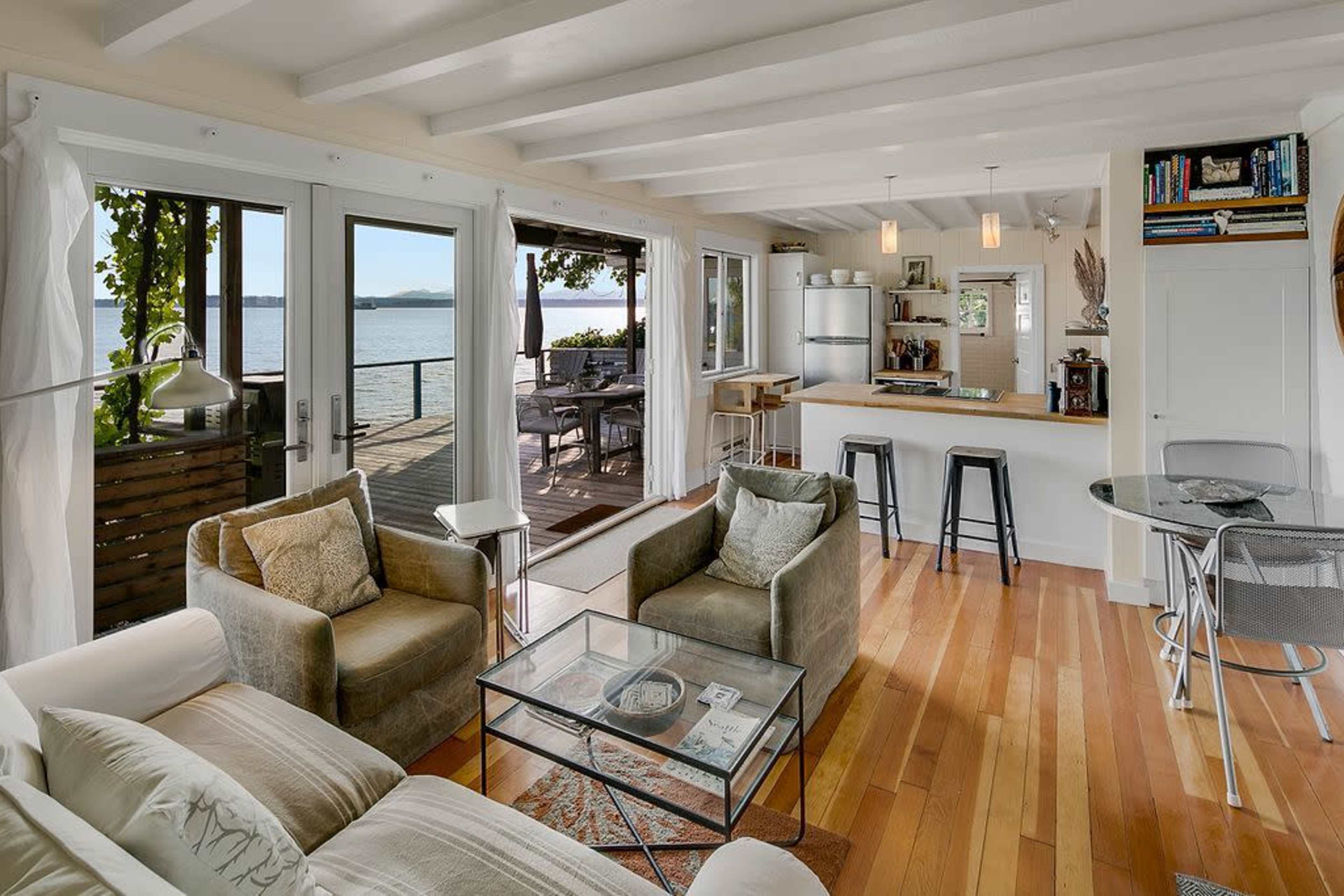 A bright living area with a view of the water, featuring a glass table, two upholstered chairs, and a kitchen bar with stools.