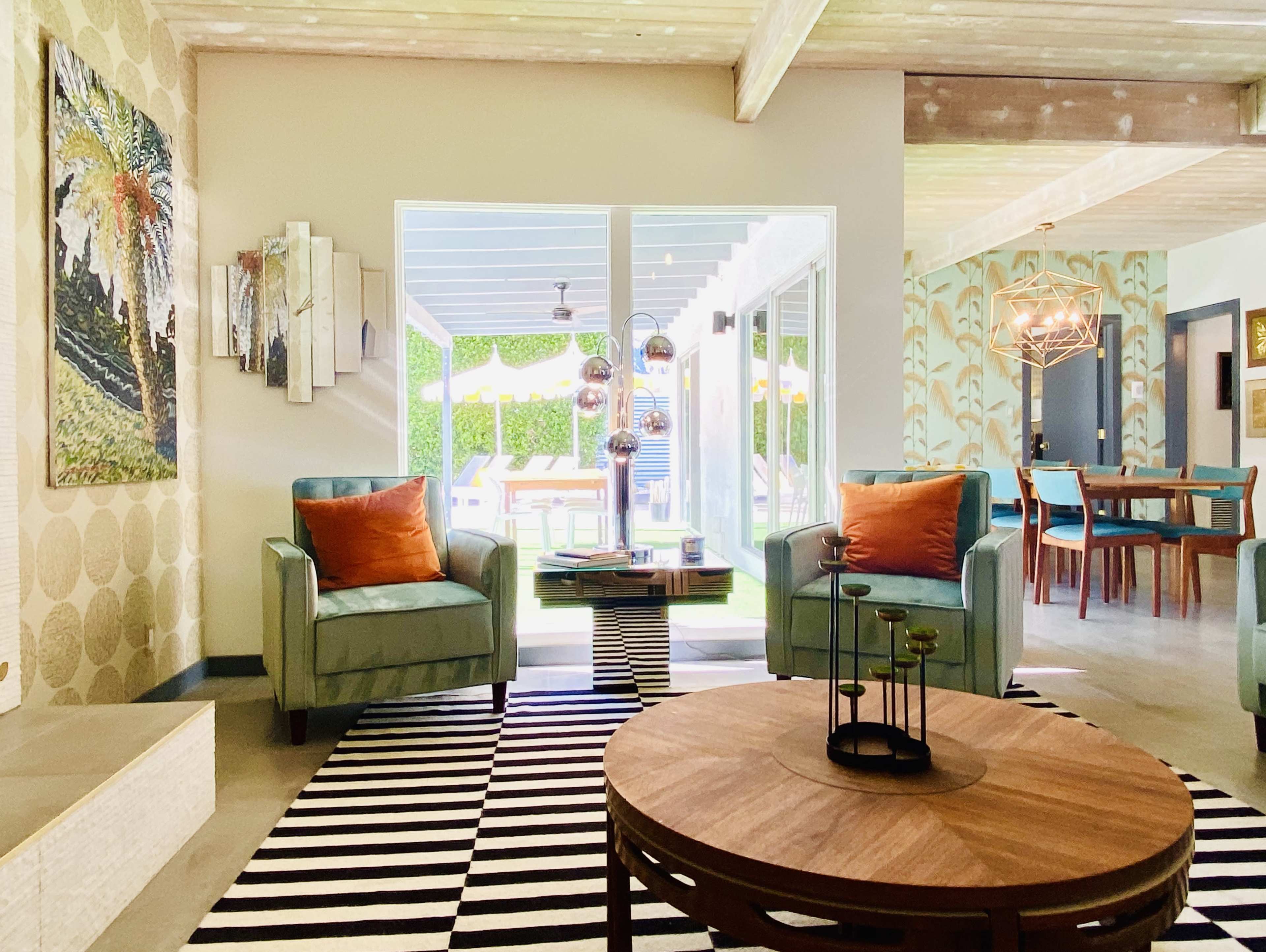 A modern living room featuring two teal armchairs with orange pillows, a round wooden coffee table, and large windows overlooking an outdoor area with patio furniture.