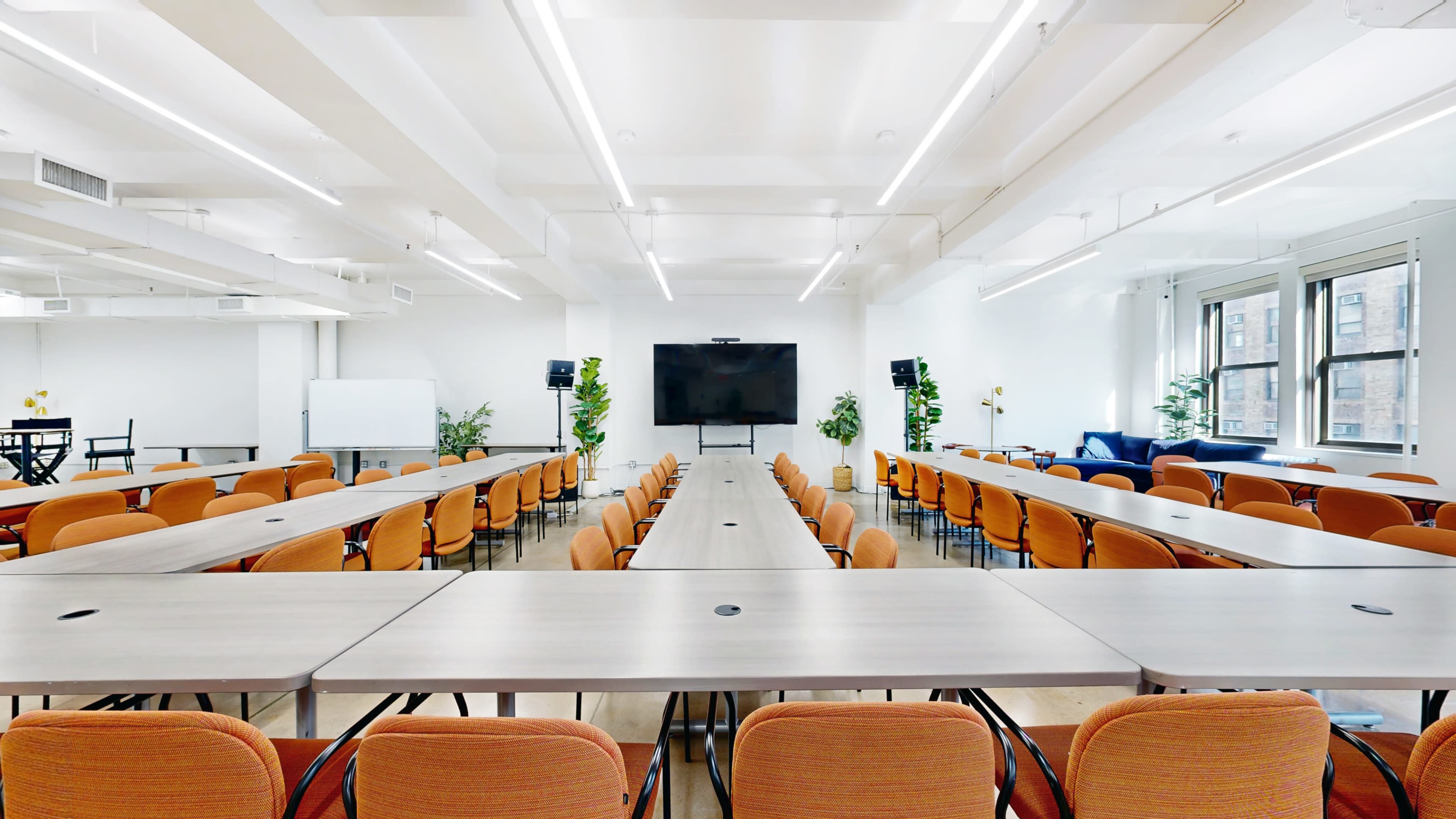 The image shows a modern conference room with arranged tables and chairs, a large screen at the front, and plants positioned around the room.