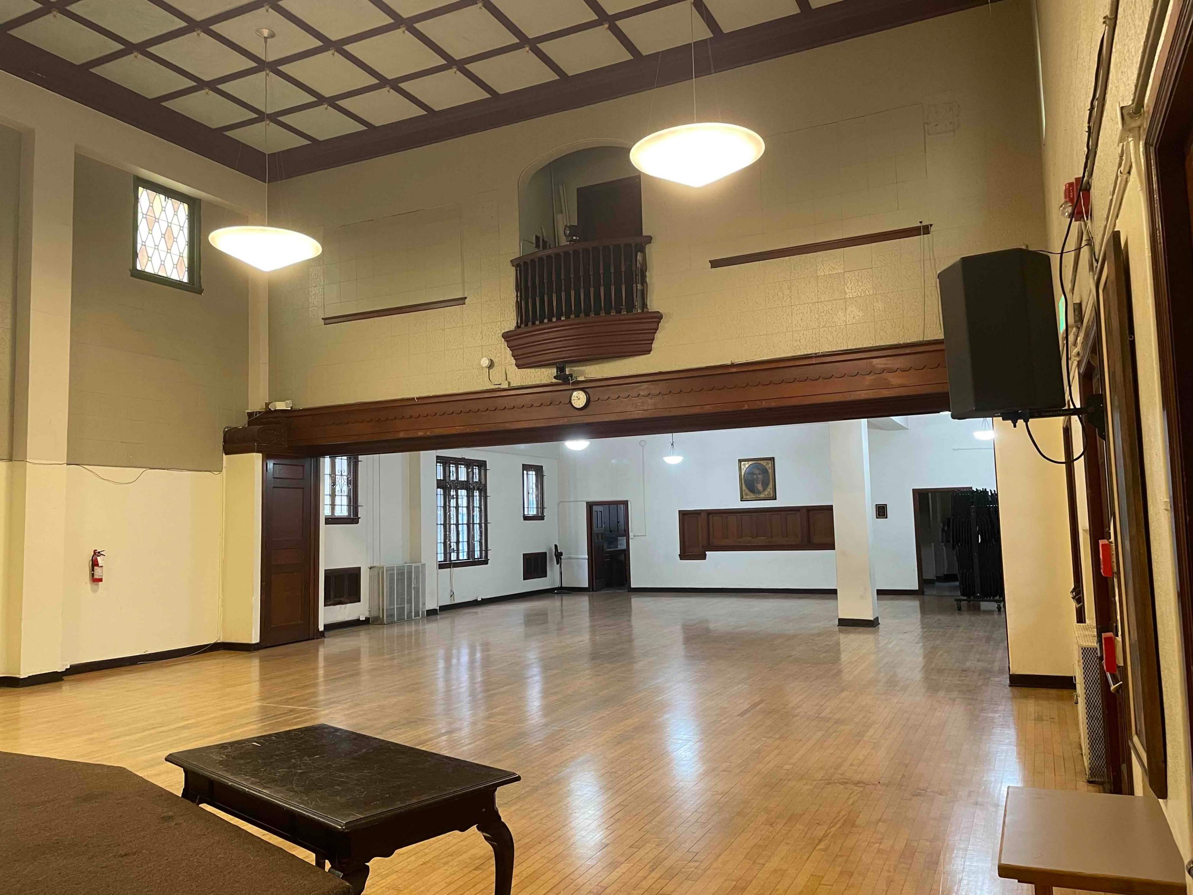 The image shows a spacious, well-lit room with wooden floors, high ceilings, and a balcony area along one wall.