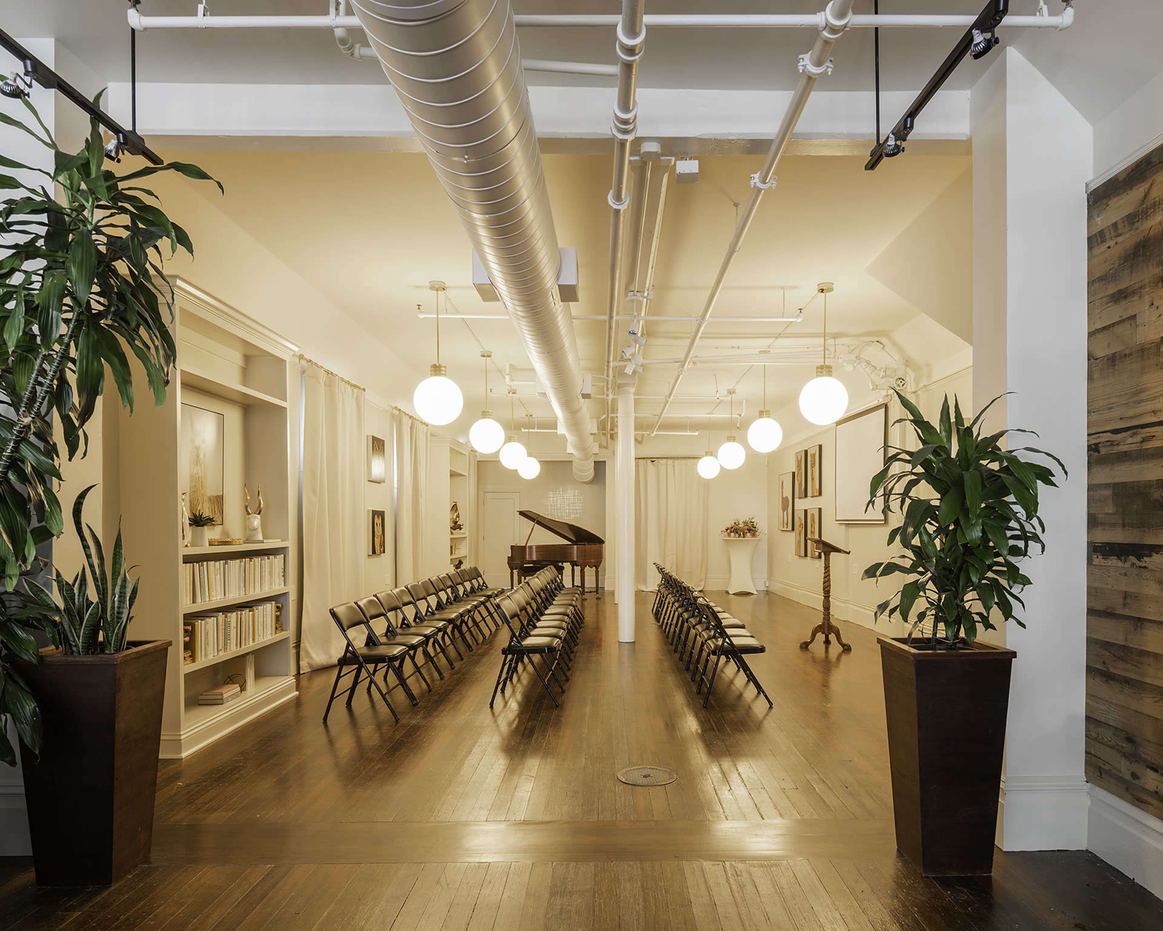 A spacious room with wooden floors is set up for an event, featuring rows of folding chairs and a piano at the front, surrounded by plants and softly lit by pendant lights.