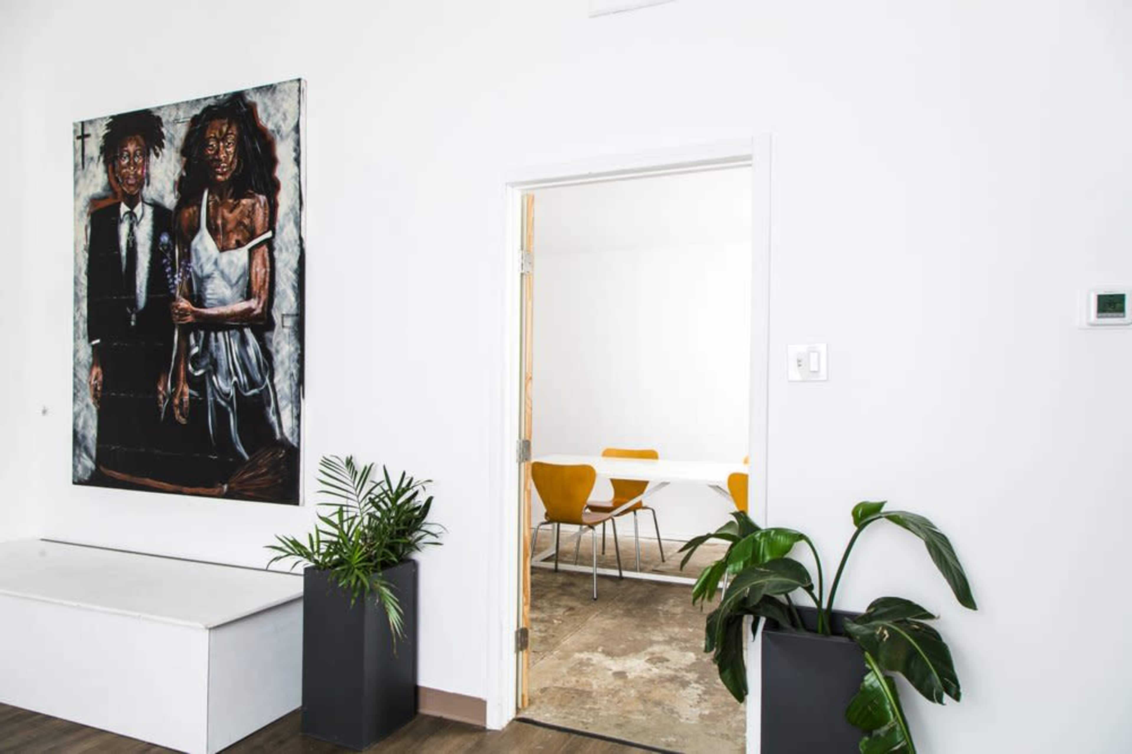 An interior space with a large painting of two figures on a white wall, a doorway leading to a bright room, and potted plants in the foreground.