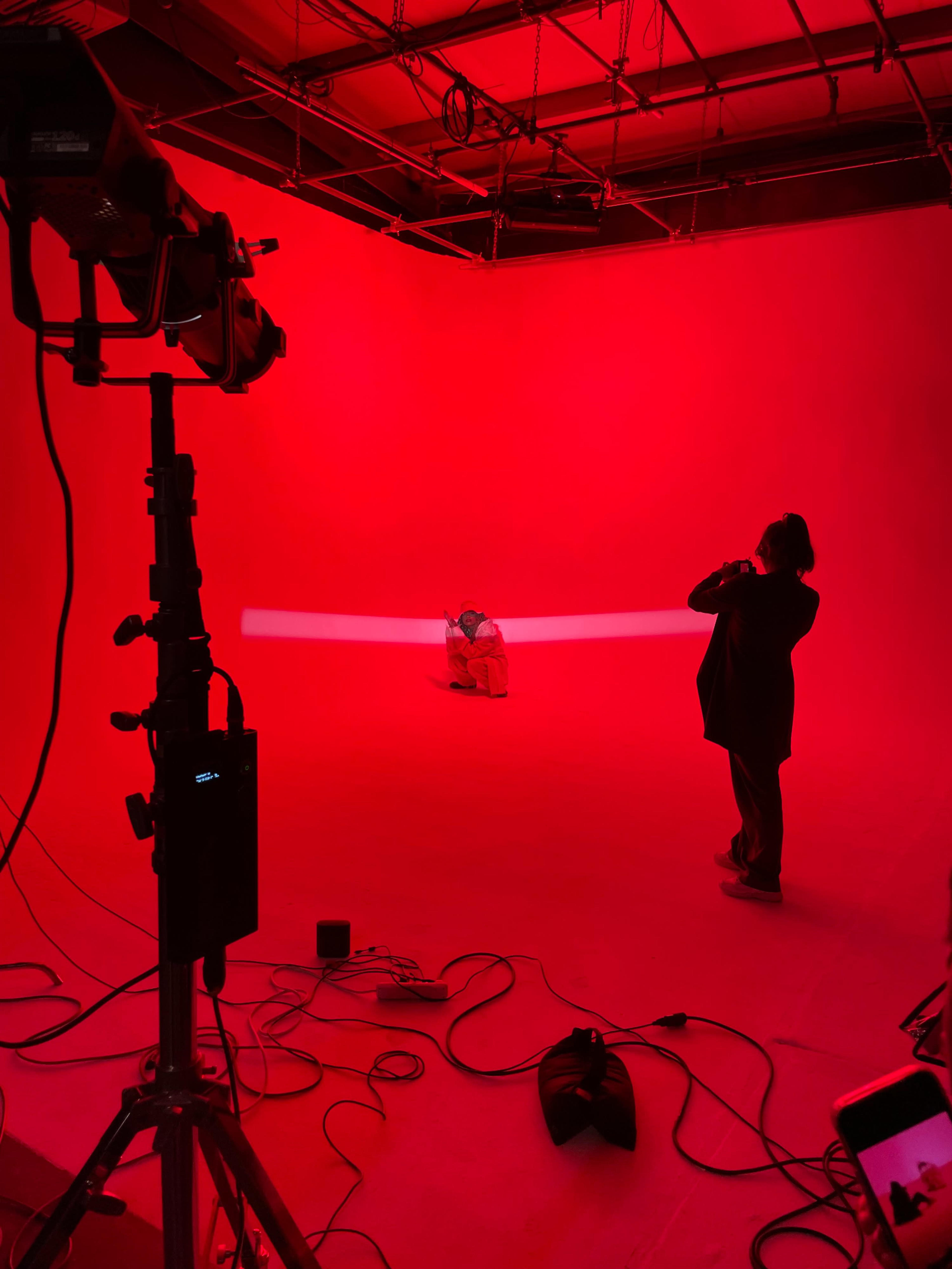 A person is taking photos of a seated model in a red-lit studio with various lighting equipment visible.