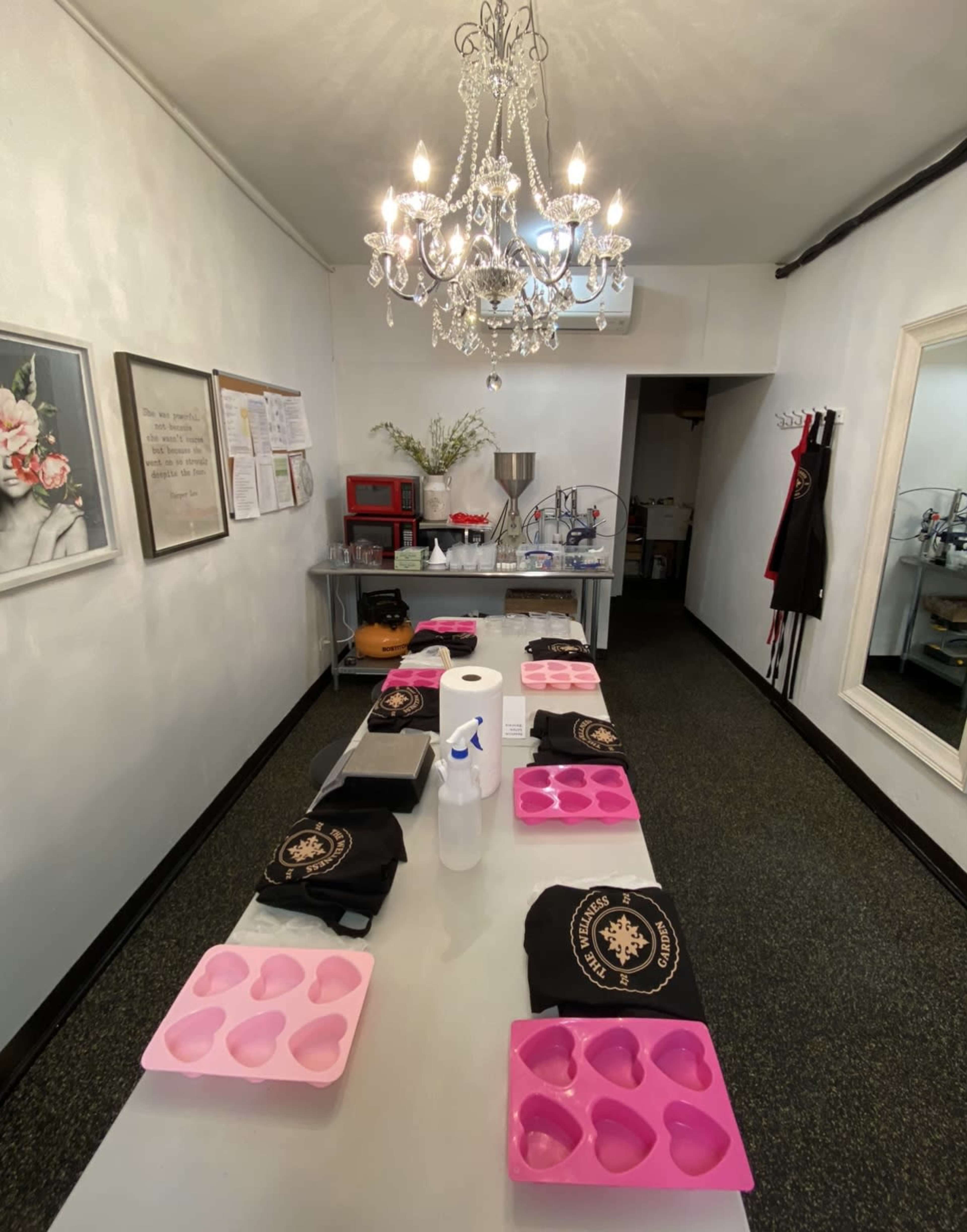 The image shows a well-lit room with a long table covered in pink heart-shaped molds, cleaning supplies, and black garments, all beneath a chandelier.