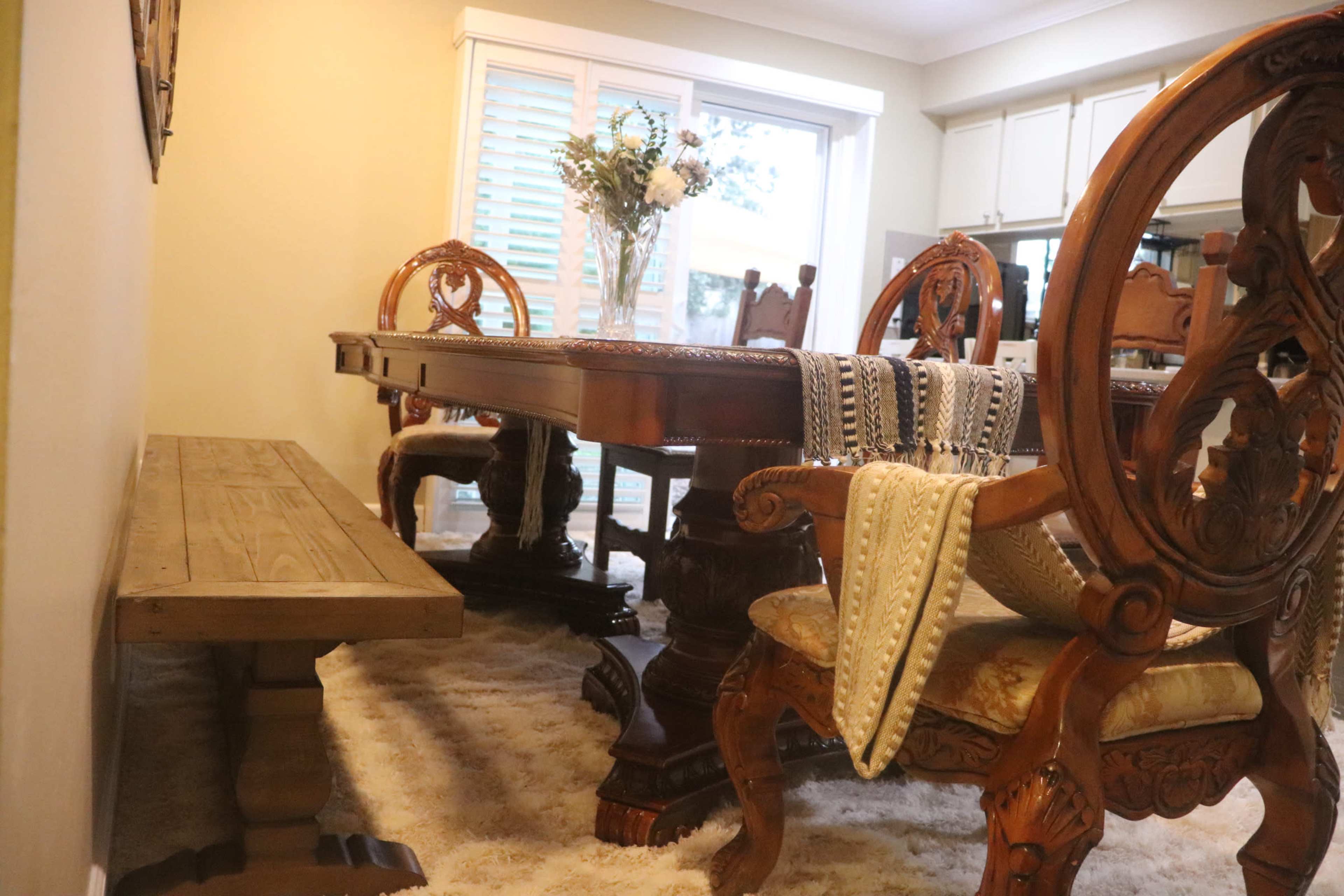A wooden dining table with intricately carved chairs is set in a brightly lit room, accompanied by a rustic bench along one side.