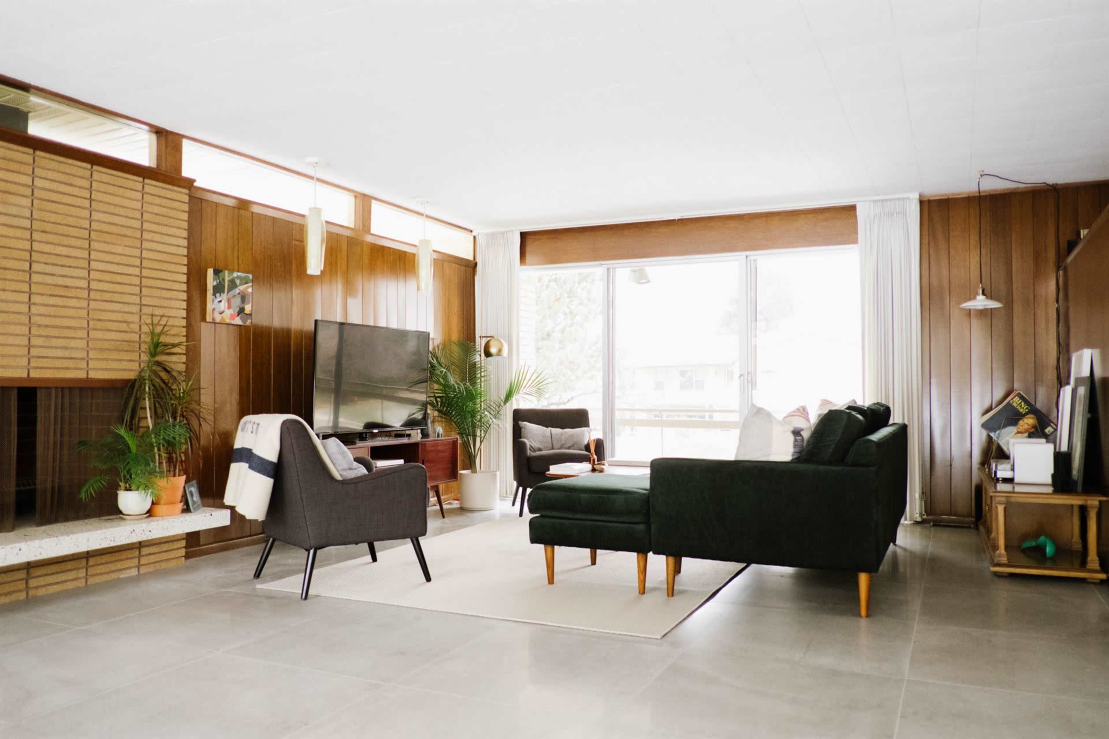 A modern living room with a dark green sectional sofa, two gray armchairs, a television, and large windows allowing natural light.